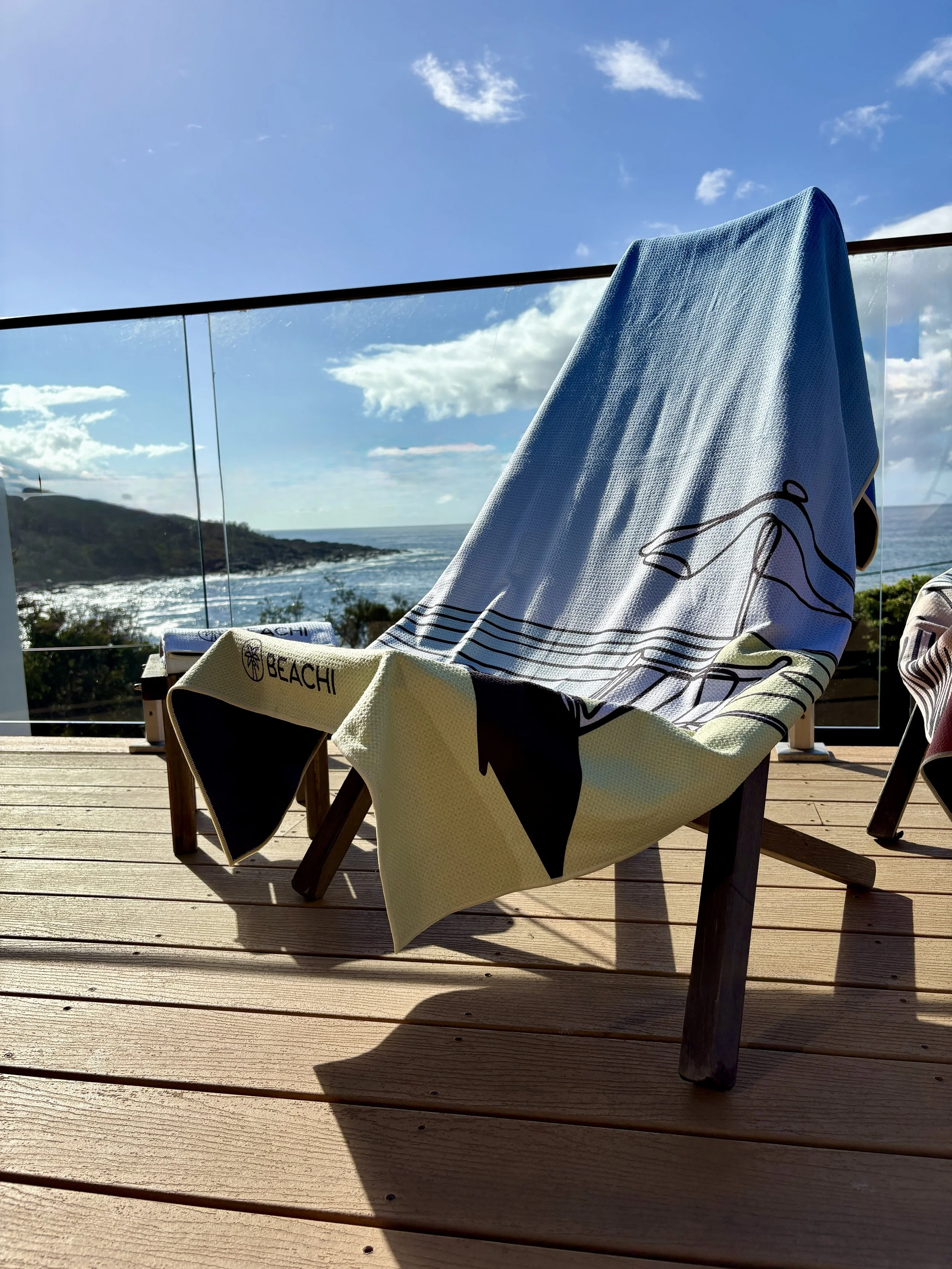 Beach chair with a Travel Ready BEACHI towel draped over it on a wooden deck, overlooking the ocean and a hillside under a partly cloudy sky.