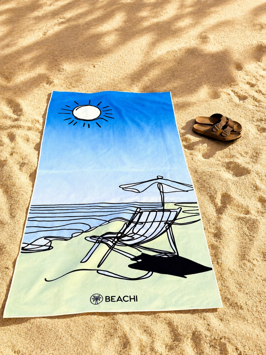 A colorful beach towel with a drawing of a chair, umbrella, and shoreline, labeled 'BEACHI,' This towel design is called 'Lazi Coast' on sandy beach next to a pair of brown sandals. This is a Sand-Free, Quick-Dry Towel