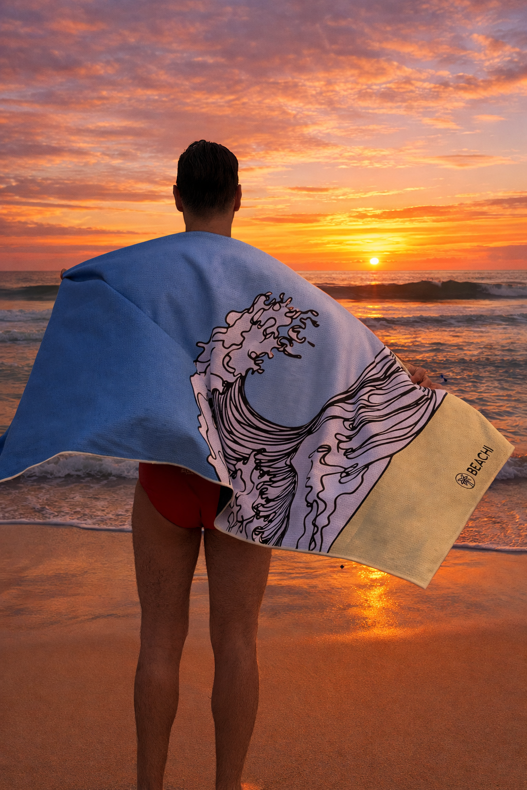 Man with short dark hair and red swim trunks standing on the beach at sunset, holding a Quick Dry BEACHI towel with a wave illustration, facing the ocean with the sun setting over the water.