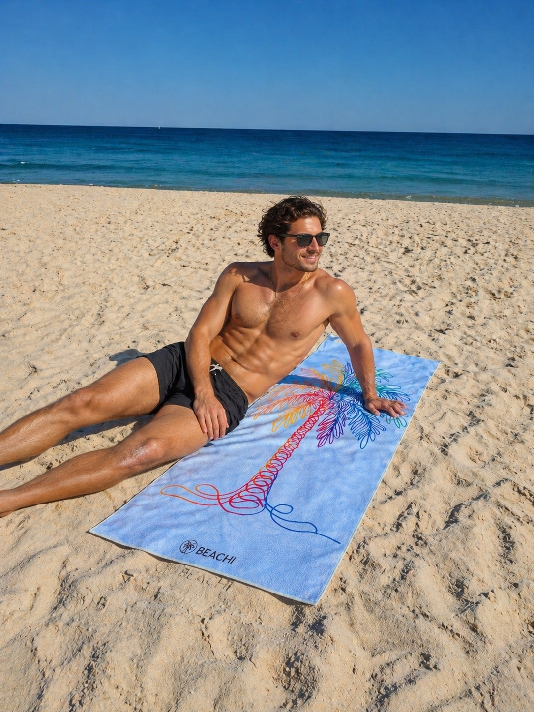 A shirtless man with sunglasses lying on the BEACHI Tropique Quick-Dry beach towel on the sand, with the ocean and sky in the background.
