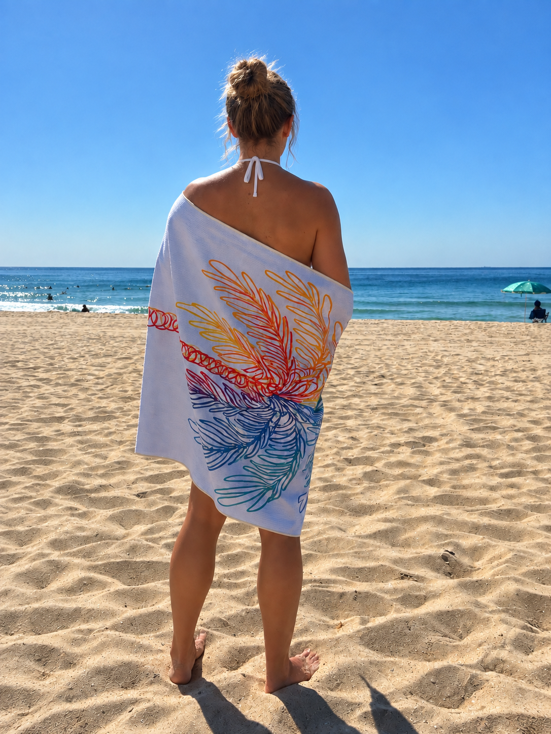 A woman standing on an Australian beach, wrapped in a colorful Beachi Quick-Dry Sand-free towel with leaf patterns, facing the ocean with clear blue skies.
