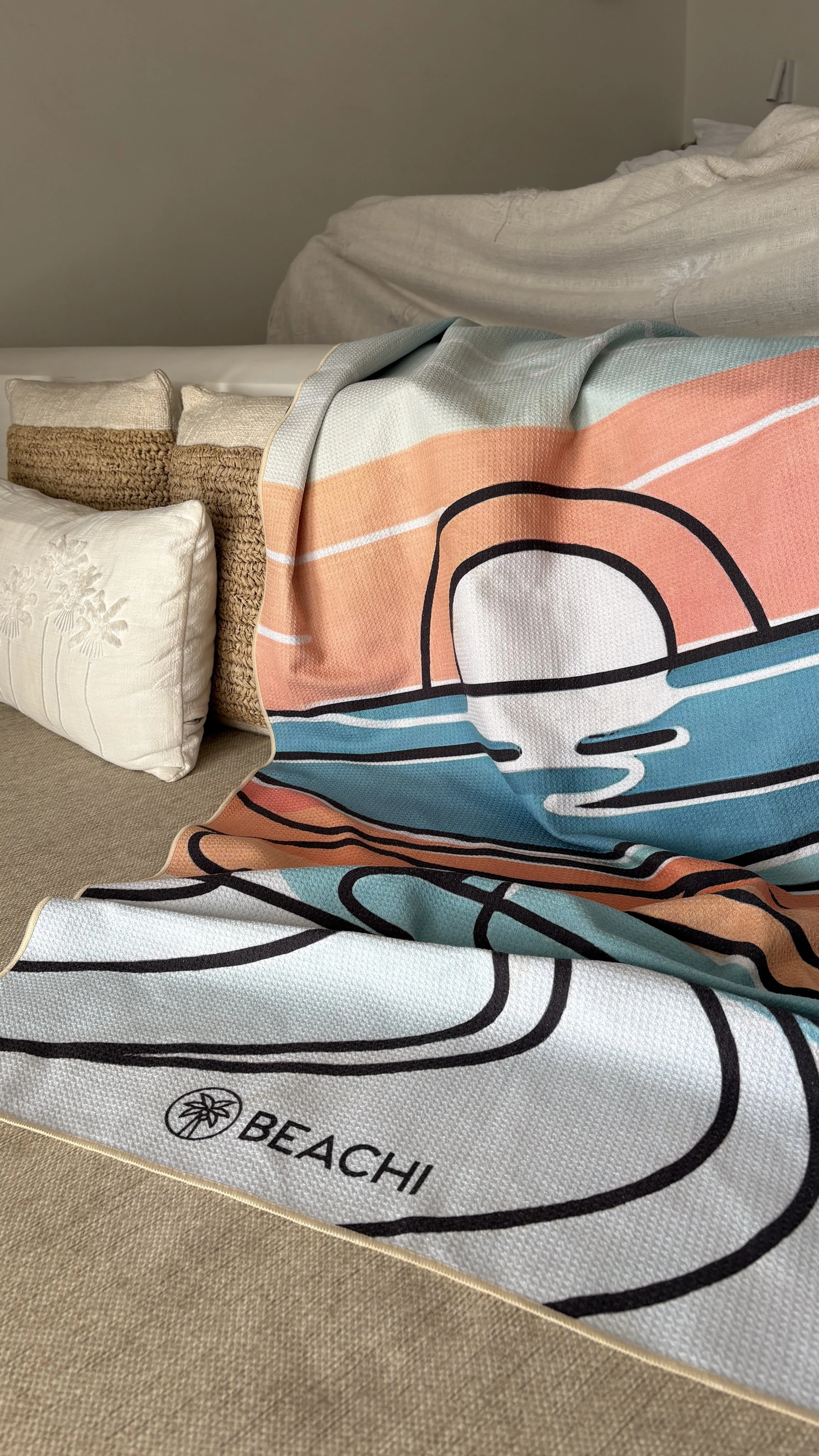 Colorful beach towel with a graphic design featuring a sunset, water, and sand draped over a bed, alongside white and beige pillows in a hotel room.