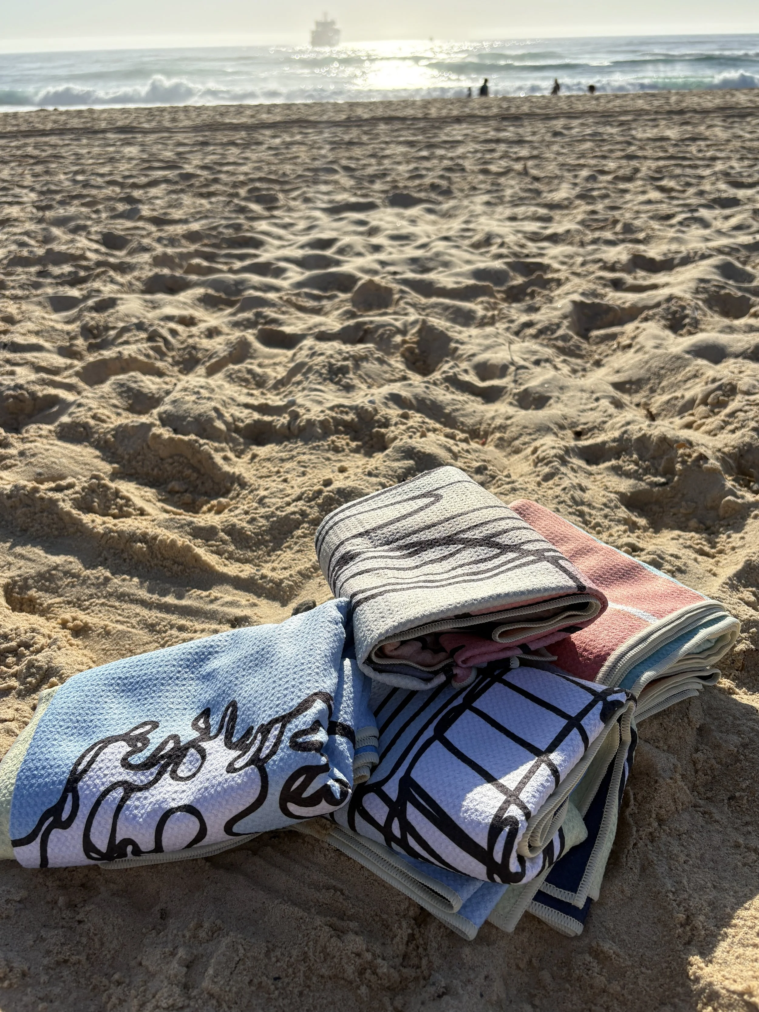 A stack of folded Sand-free towels on sandy beach with ocean waves and a ship in the distance under a bright sun.