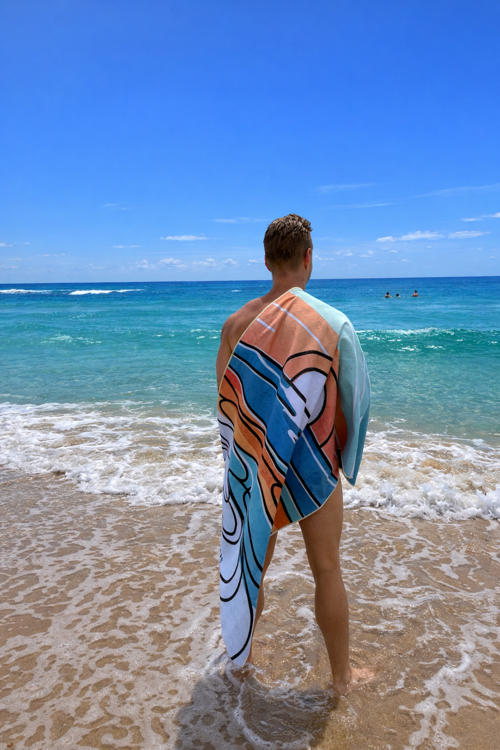 A person standing at the beach with a colorful Sand-Free BEACHI towel draped over their shoulders, looking out at the ocean under a clear blue sky.