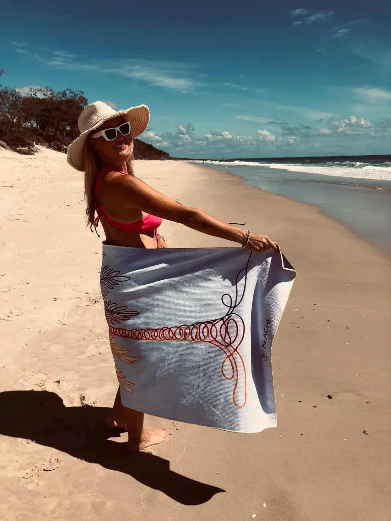 A woman standing on a sandy beach with ocean waves in the background, wearing a large straw hat, white sunglasses, a red bikini top, and holding a large striped Quick Dry, Sand-Free BEACHI towel.