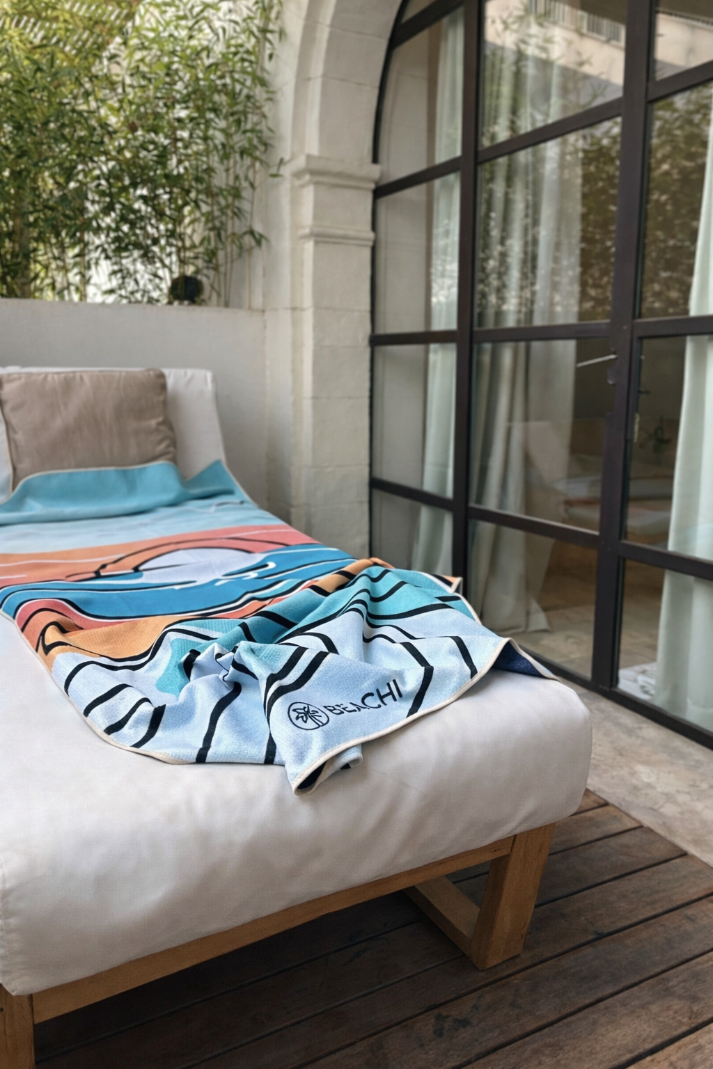 An outdoor daybed with beige cushions and a multicolored beach towel featuring the word "BEACH" and a palm tree logo, set on a wooden deck beside a glass windowed wall with curtains, with greenery visible in the background.