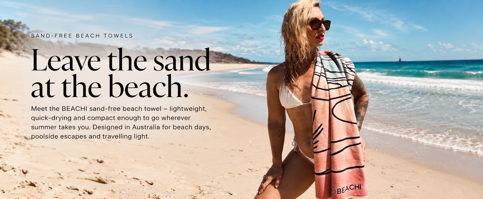 A woman with blonde curly hair, wearing sunglasses and a white bikini, standing on a sandy beach with the ocean and blue sky in the background. She has a pink towel with black lines draped over her shoulder and is looking into the distance.