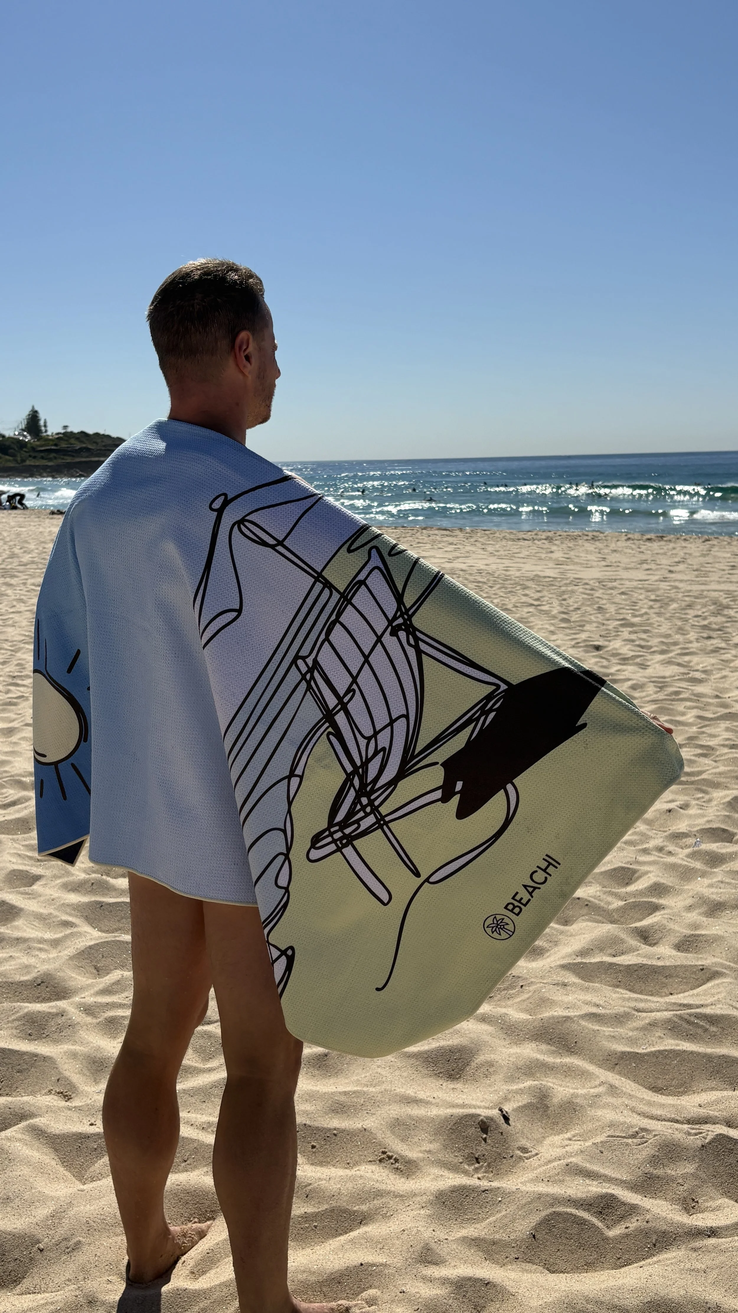 Man at a beach in Australia holding a Beachi Quick-dry Sand-free towel, looking at the ocean under a clear blue sky.