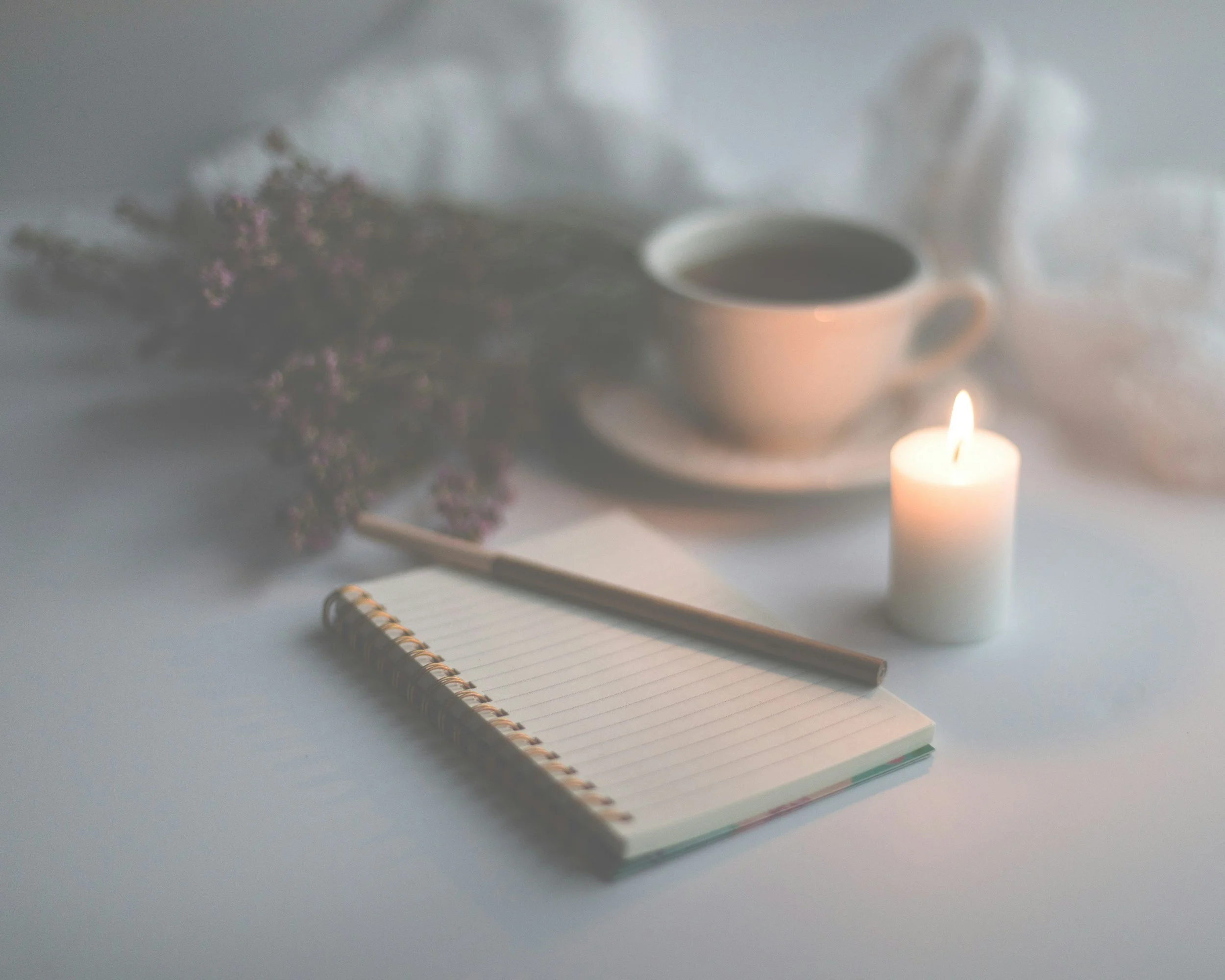 A cozy scene with a notebook, pen, lit candle, cup of coffee, and a bouquet of purple flowers on a white surface, with a white cloth in the background.
