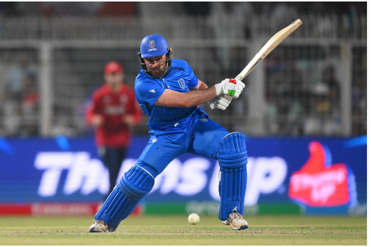 A cricket player in blue uniform and protective gear is batting on the field with a cricket bat. An opponent in a red uniform is in the background.