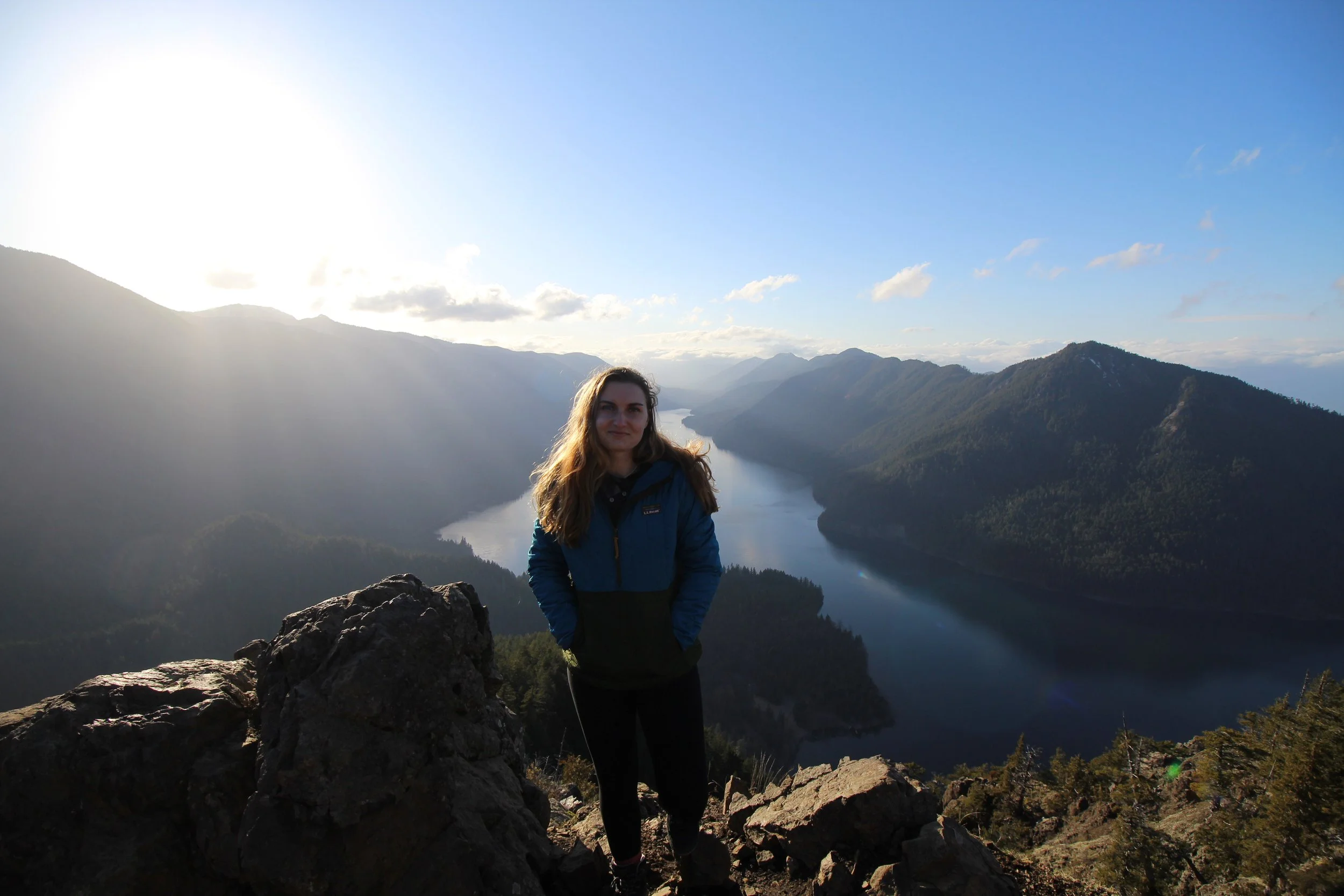 Chelsea standing on a rocky cliff with a scenic view of a river and mountains in the background during daylight, located on the Olympic Peninsula in Washington.