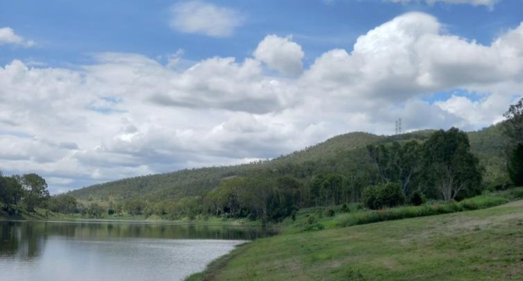 Scenic landscape with river, grassy riverbank, forested hills, and cloudy sky.