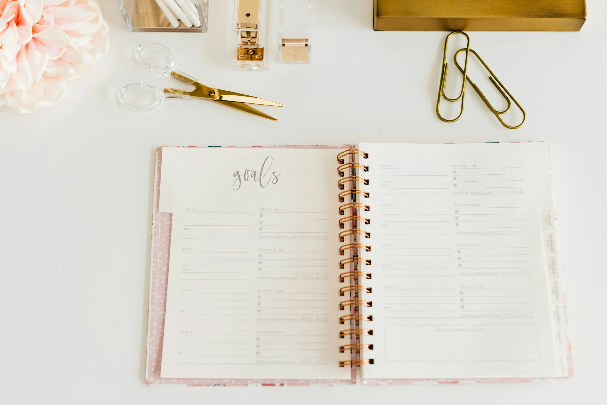 An open planner with goals and action plan pages on a white desk. Office supplies include gold scissors, gold paper clips, clear stamp blocks, and a flower arrangement.
