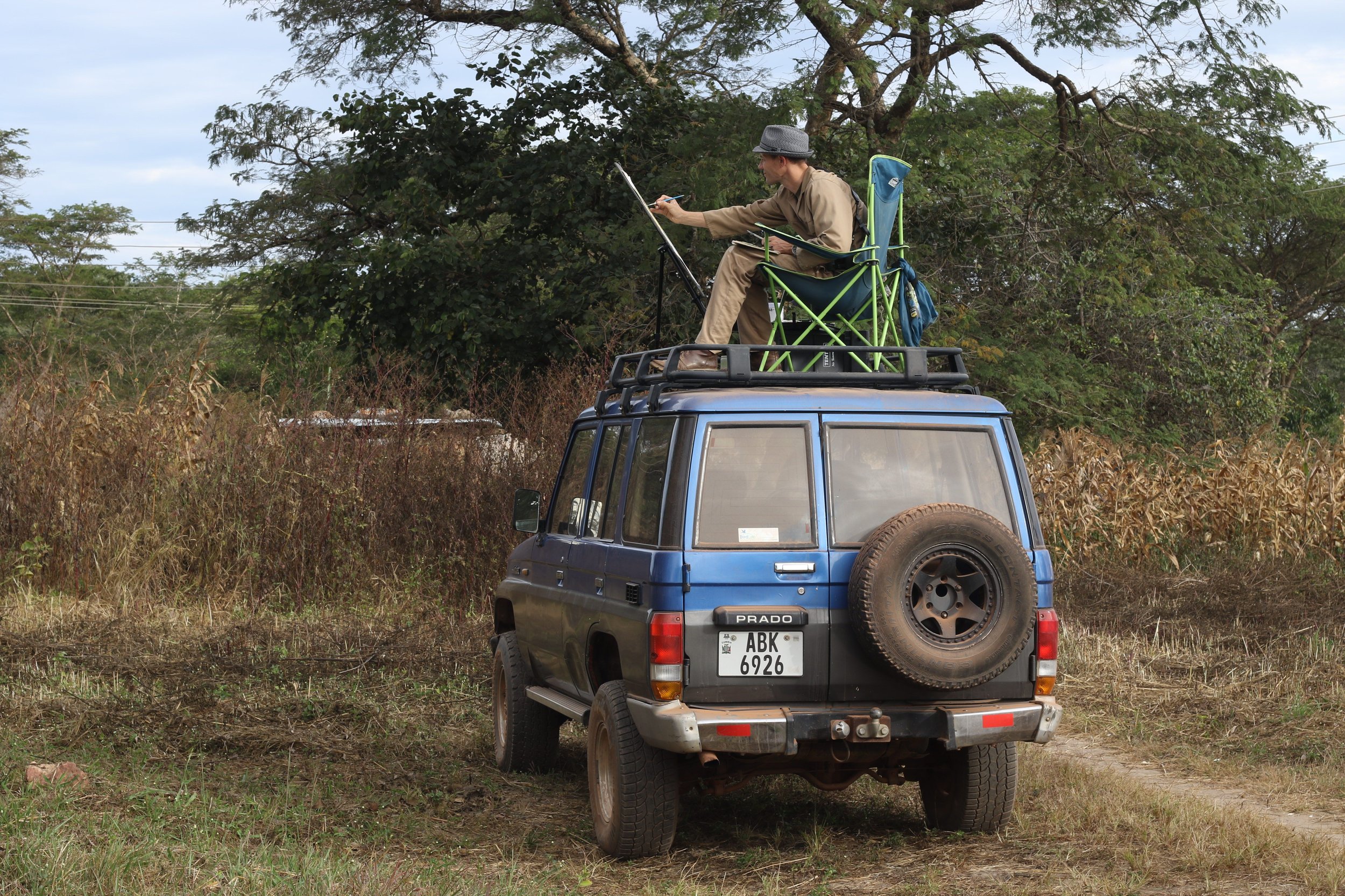 A person sitting in a chair on the roof of a parked blue SUV, painting a scene outdoors with a rural background.