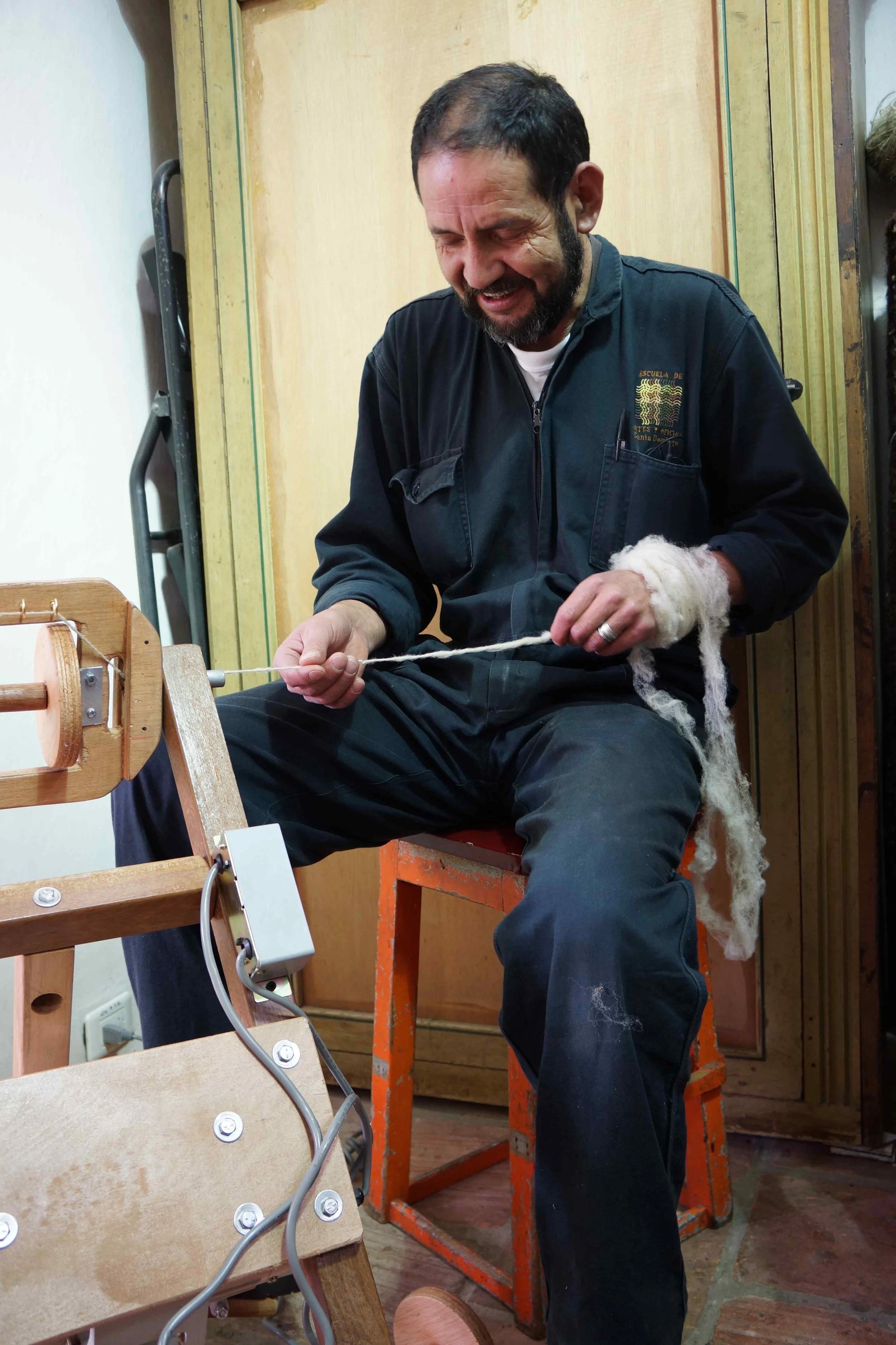 A man with a beard and dark hair, wearing a black jacket, sitting on a red-orange stool, and spinning wool into yarn using a spinning wheel, smiling while working.