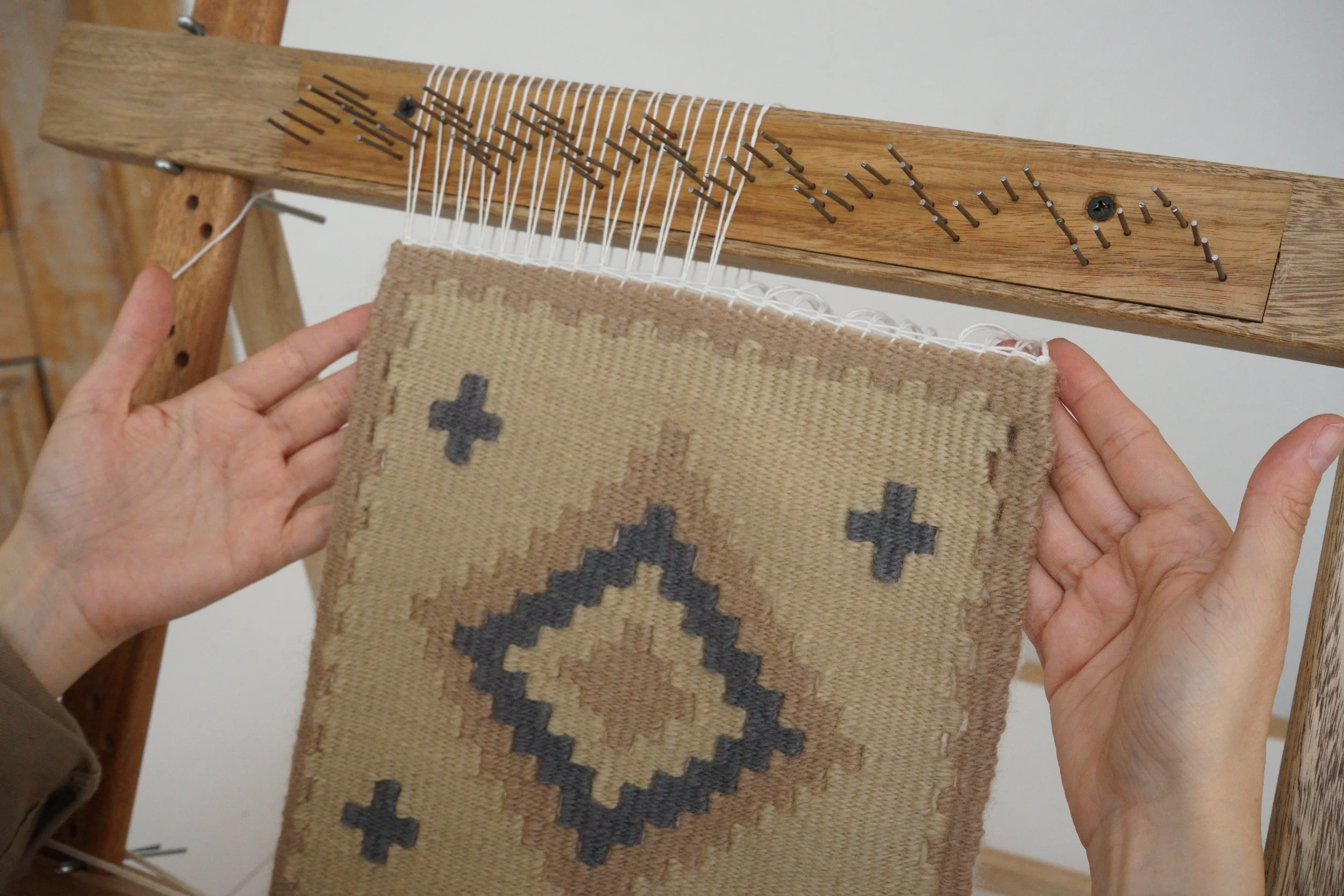 Person weaving fabric on a wooden loom, holding a weaving tool, with a piece of woven cloth on the loom.