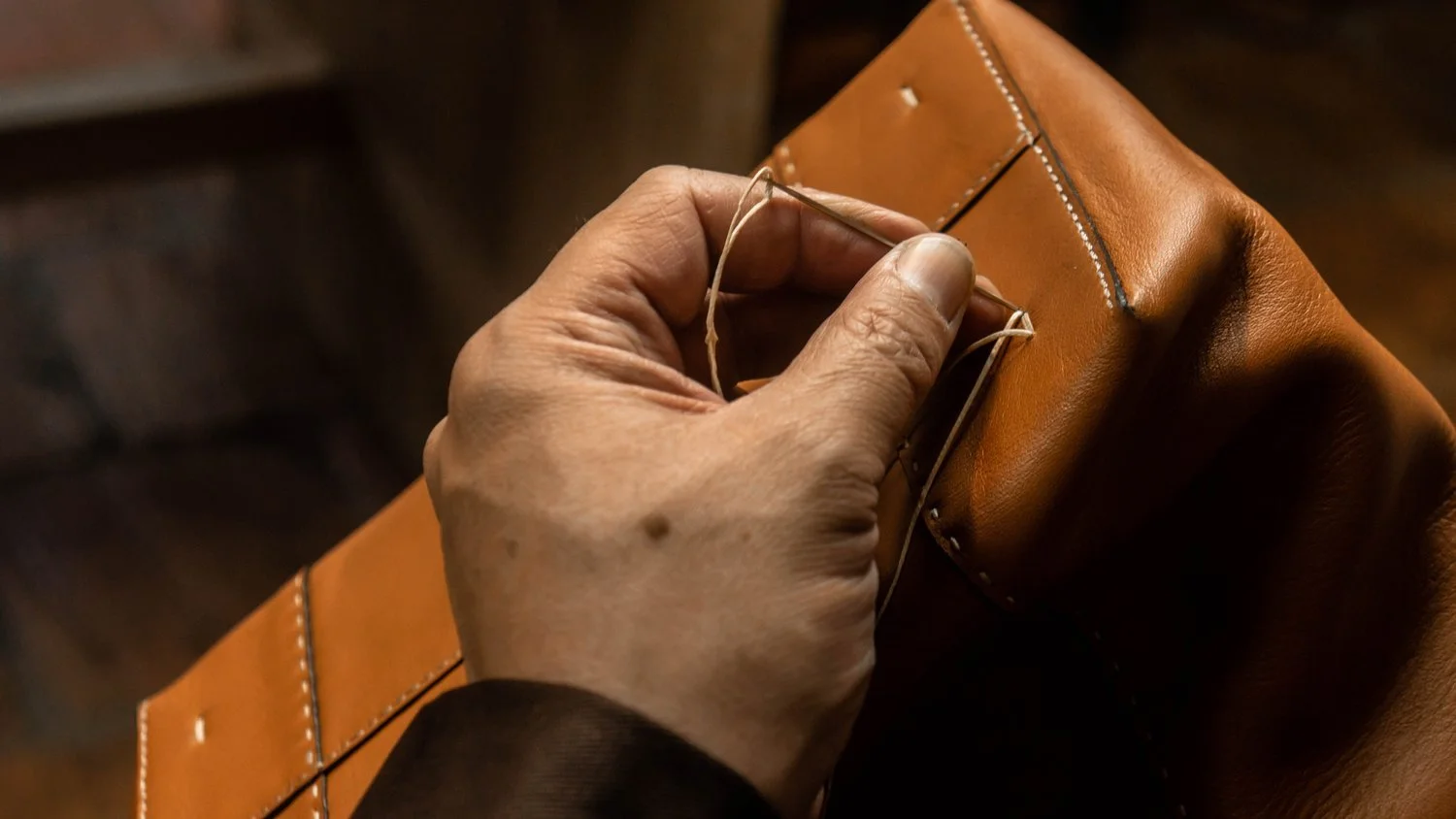 Close-up of a person hand sewing with a needle and thread on a tan leather object.