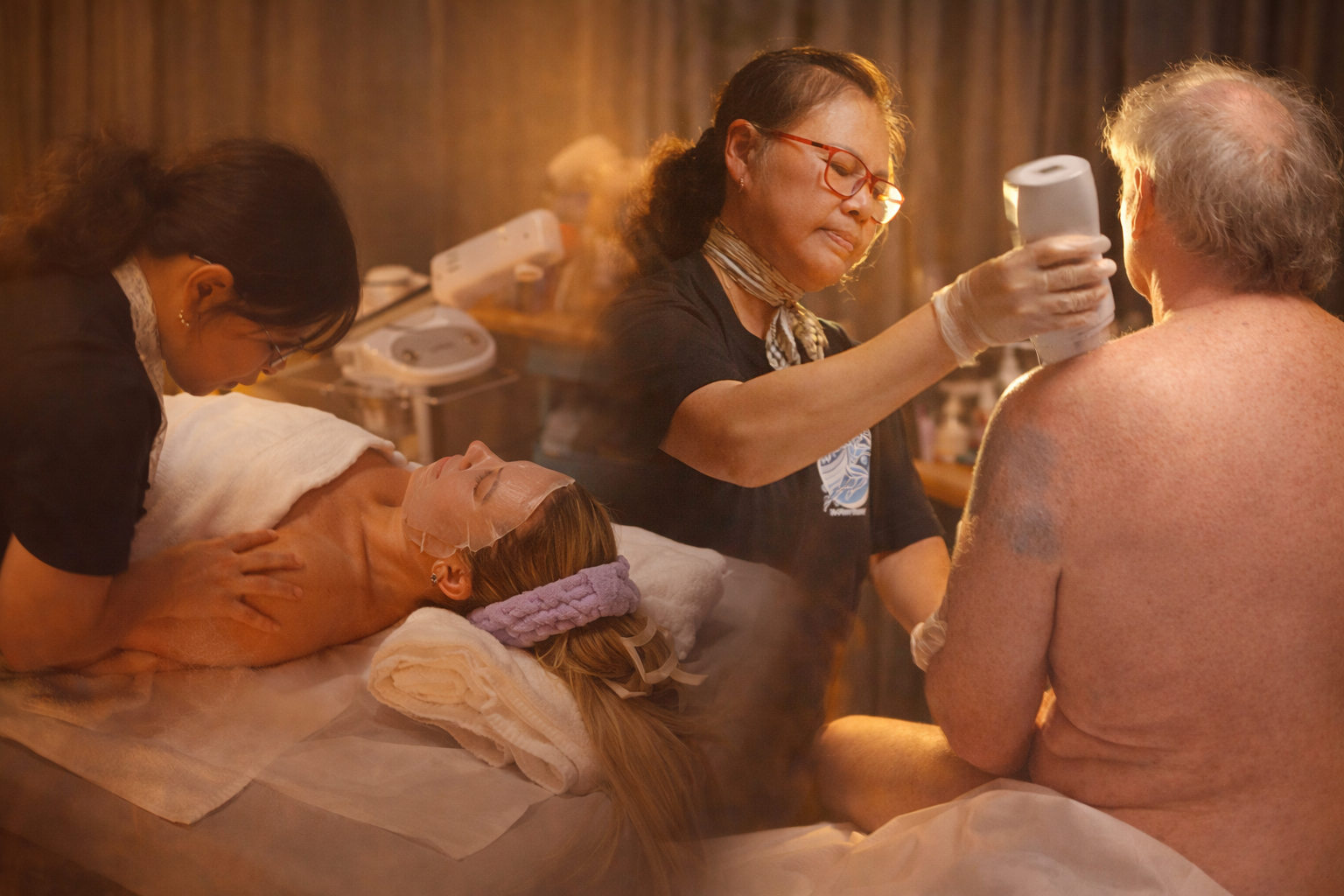 A person lying on a massage table with a facial mask, receiving a facial treatment from Mary, and Mary applies a waxing service to an older man.