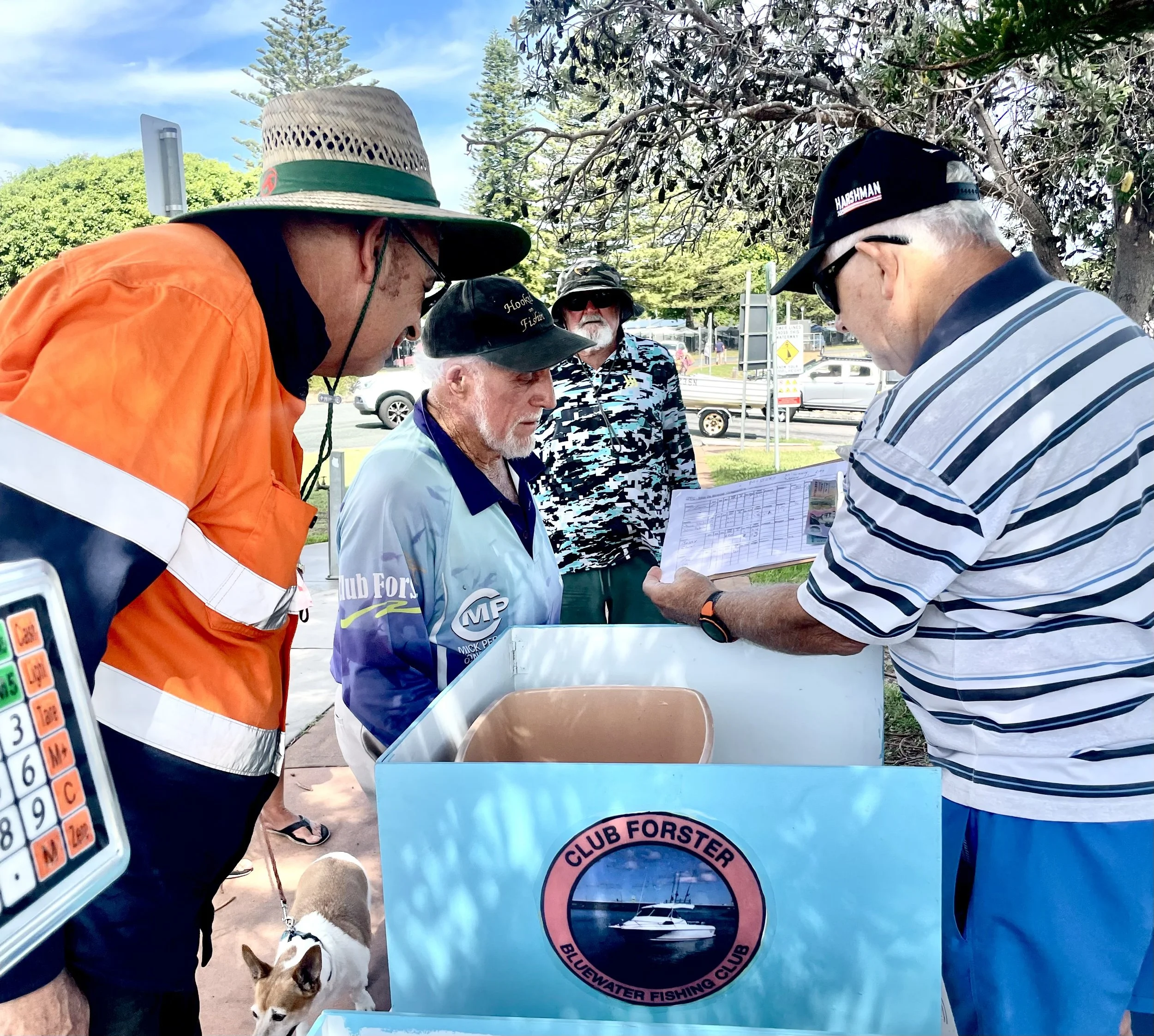 A small group of people outdoors engaging in conversation around a table. One person is holding a paper with text. There's a blue sign on the table for Club Forster Bluewater Fishing Club. One person wears a bright orange jacket, another wears a stri