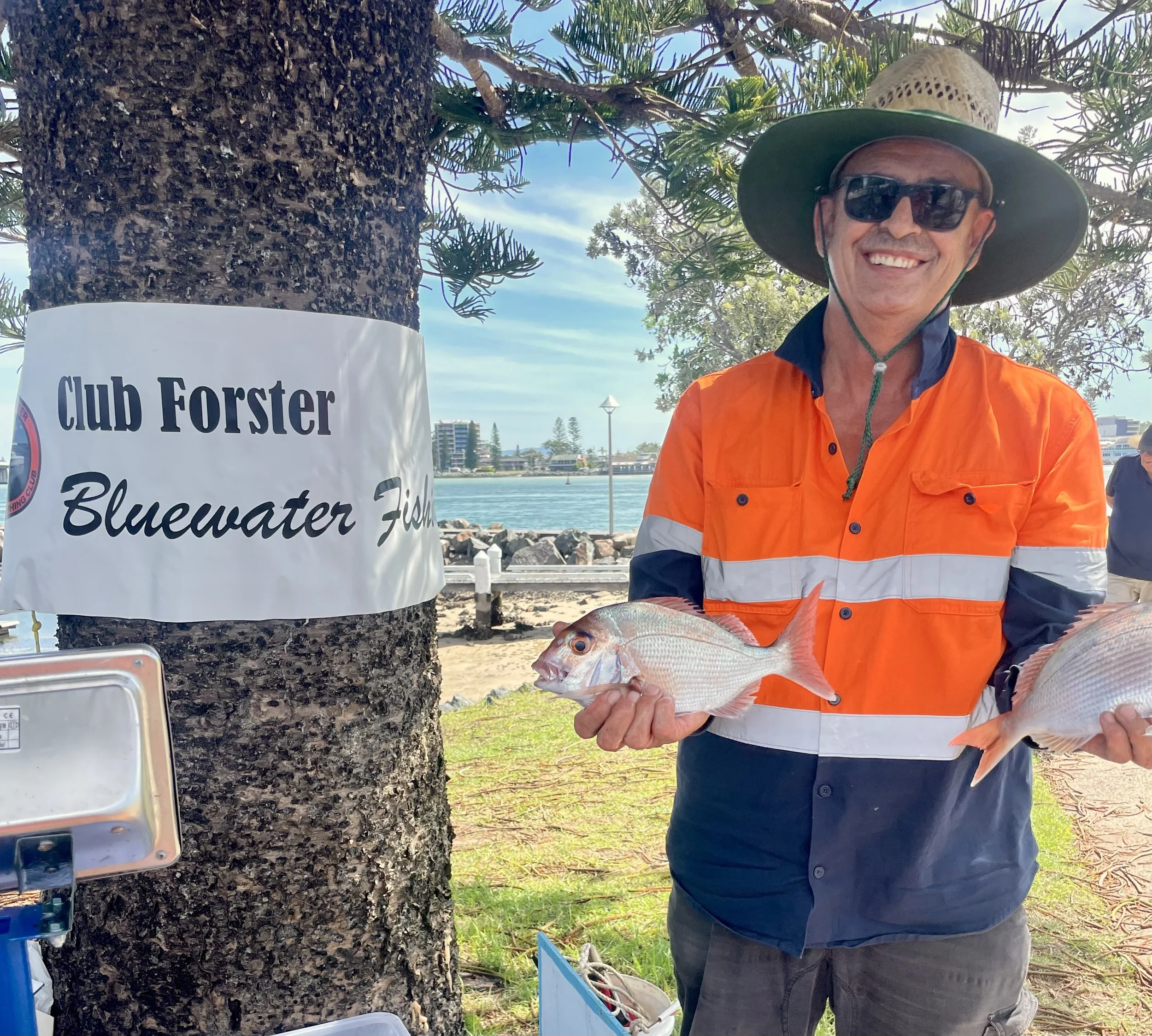 A person standing next to a tree with a "Club Forster Bluewater Fish" sign, holding two fish in their hands, wearing a bright orange and navy shirt, sunglasses, and a wide-brimmed hat. There is a body of water and buildings in the background.