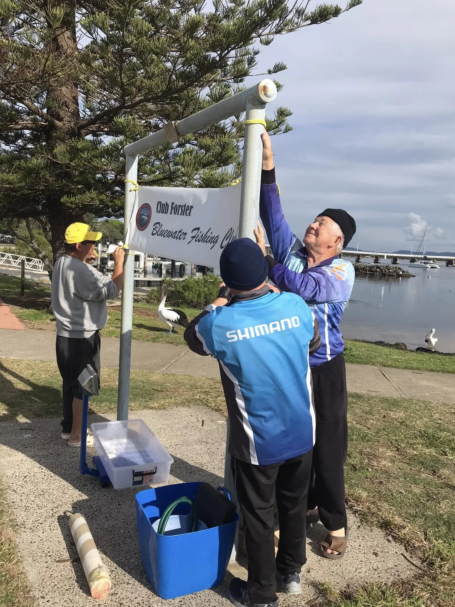 Three people setting up a sign for Club Forster Bluewater Fishing Club outdoors, near a waterfront with birds in the background.