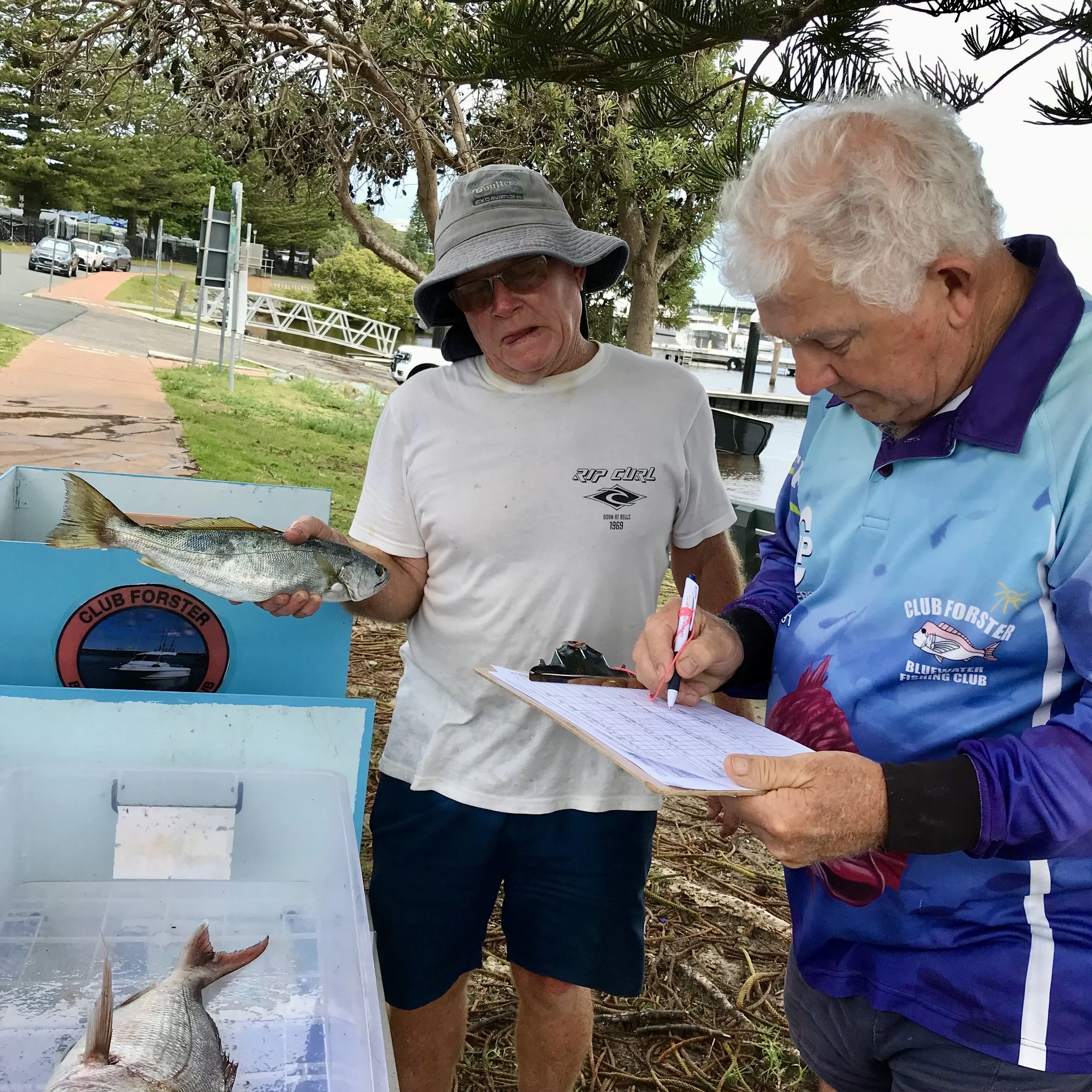 Two men by a plastic container, one holding a fish, the other writing on a clipboard.