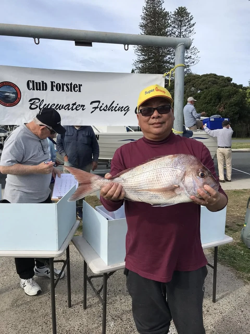 Man holding a large fish at a fishing competition, standing in front of a banner that reads 'Club Forster Bluewater Fishing.' Other people are present at the event.