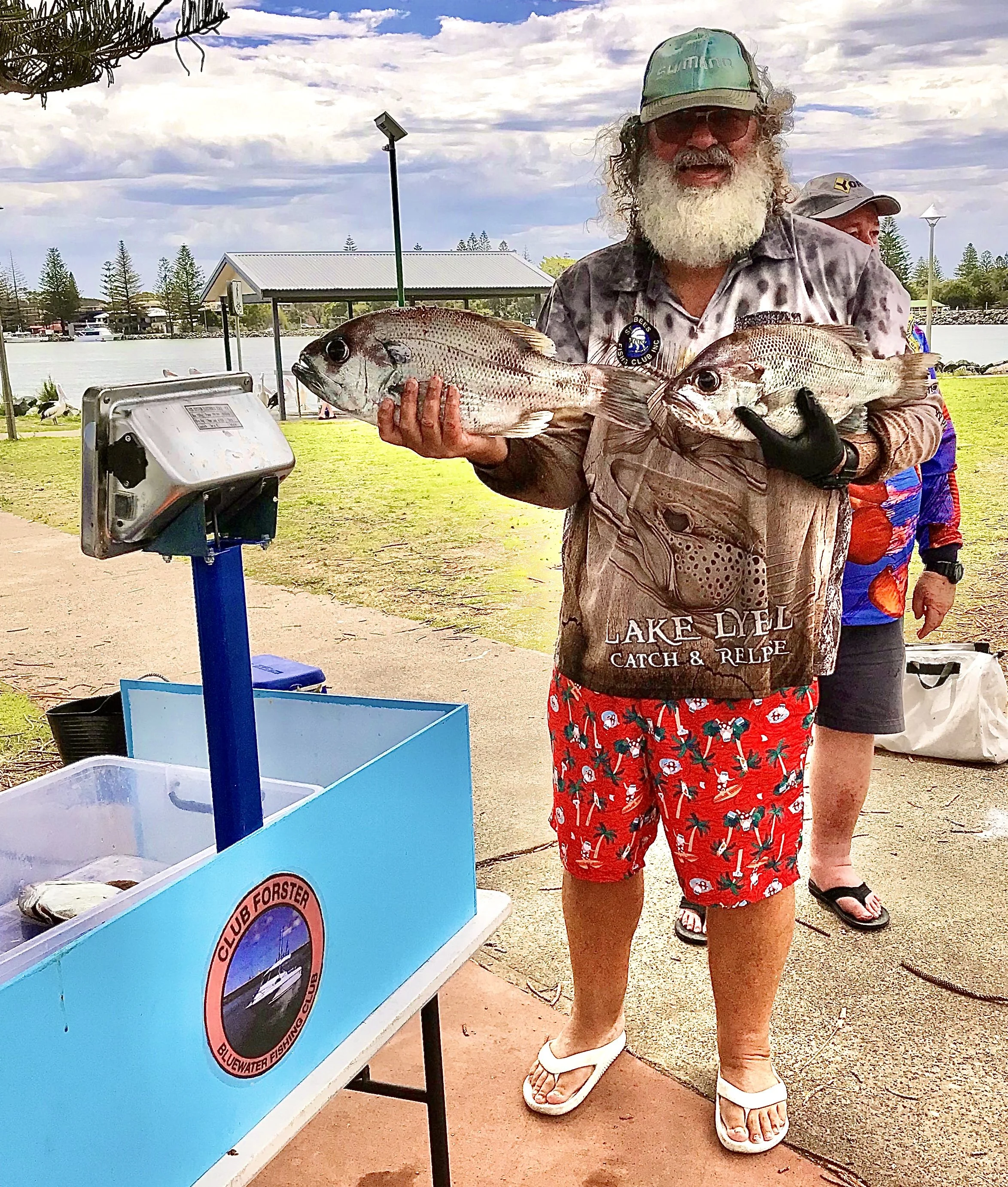 Man holding fish next to a weighing station outdoors, with "Lake Lyell Catch & Release" shirt, wearing a cap and shorts, and a background of water and trees.