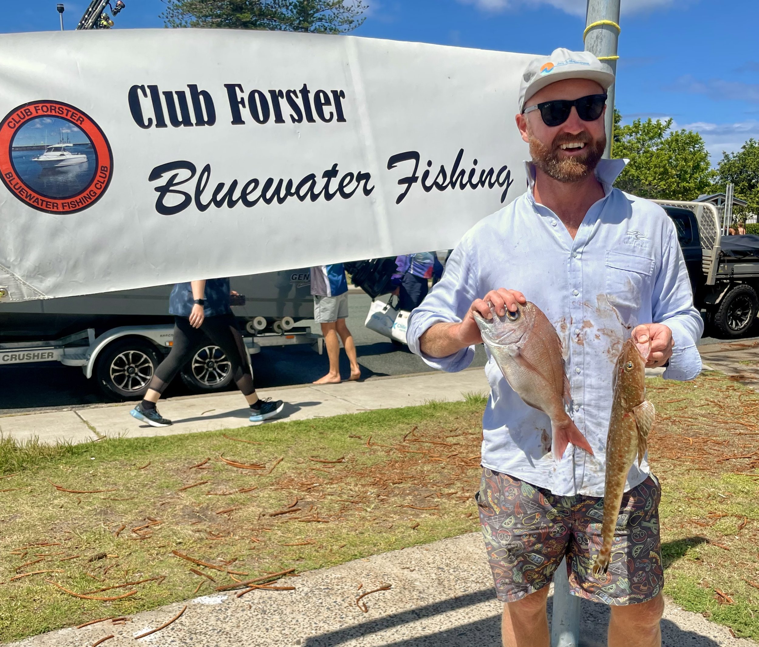 Man holding two fish at a fishing contest with "Club Forster Bluewater Fishing" banner in the background.