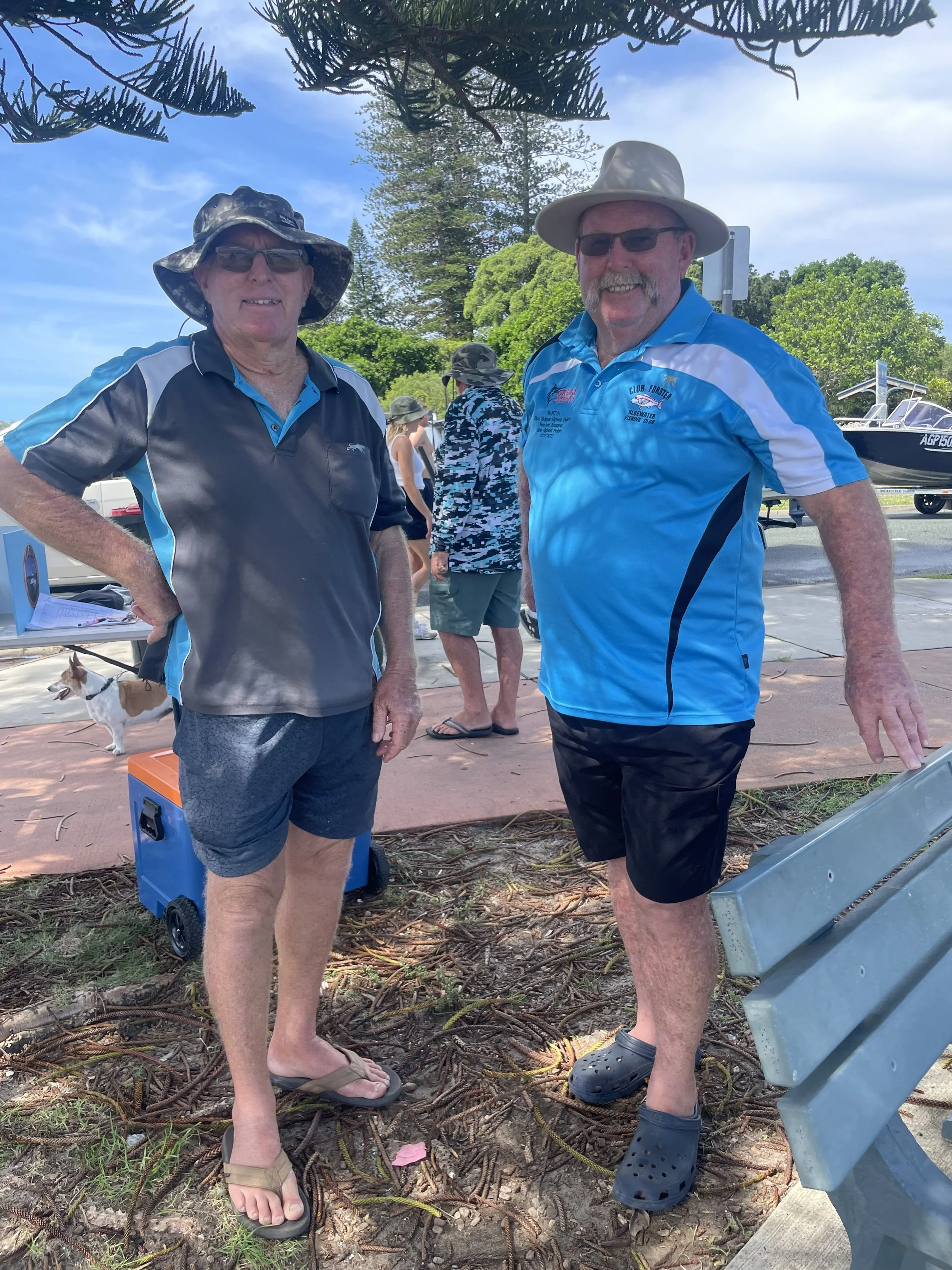 Two men wearing hats, casual clothing, and sunglasses standing outside near a bench, with trees and a boat in the background.