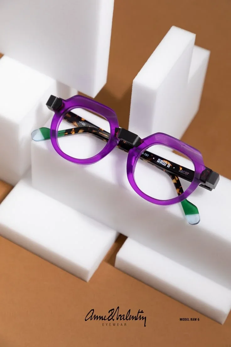 ýrazné brýle Anne et Valentin, model RAW 6, položené na minimalistických bílých podstavcích. Průsvitný fialový acetát dodává siluetě obruby lehkost a kultivovaný výraz.