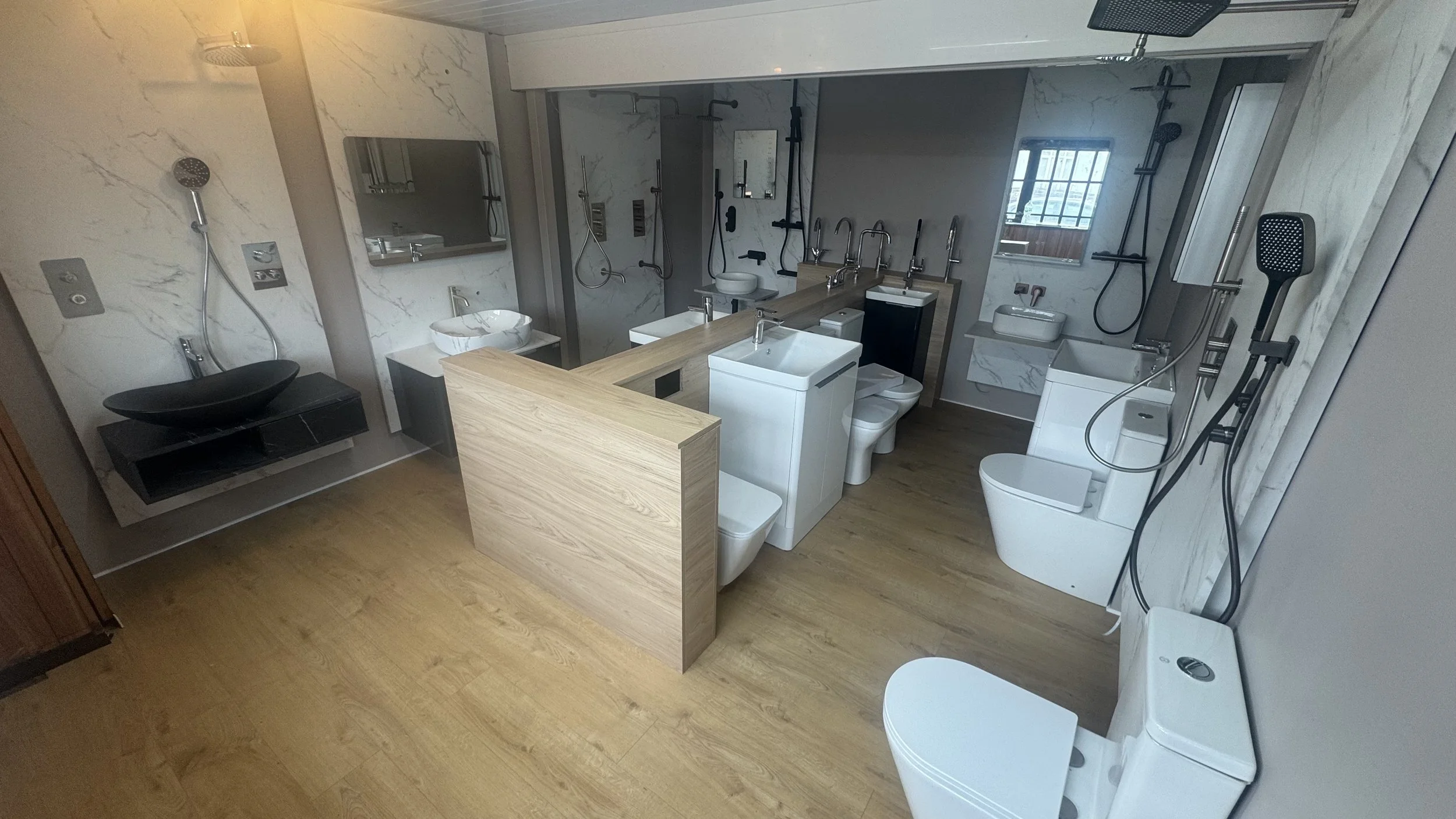 Showroom with various modern bathroom fixtures including sinks, toilets, and showerheads installed on marble walls and wooden floors.
