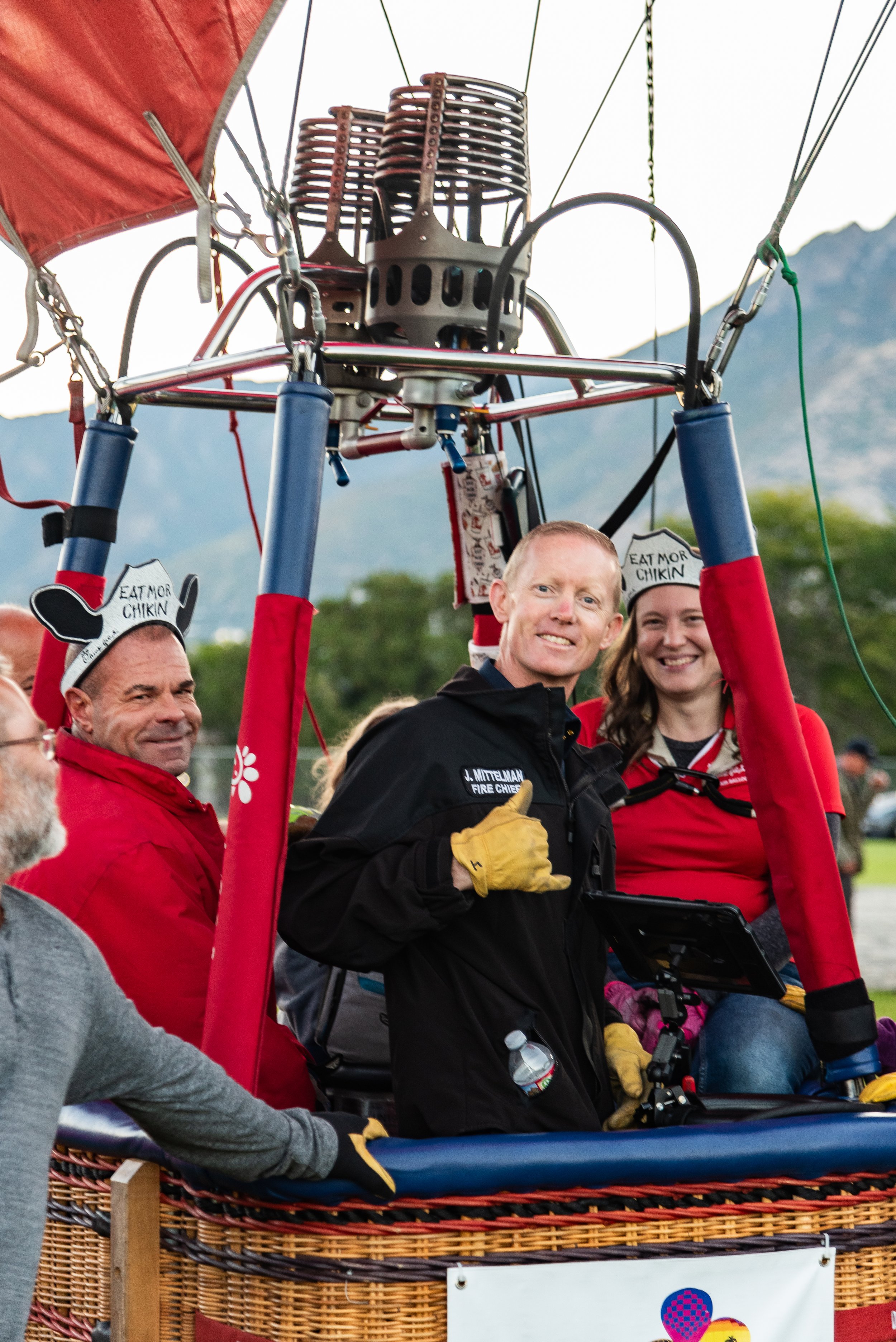 People inside a hot air balloon basket preparing for a flight, with one wearing a novelty hat and another in a uniform.