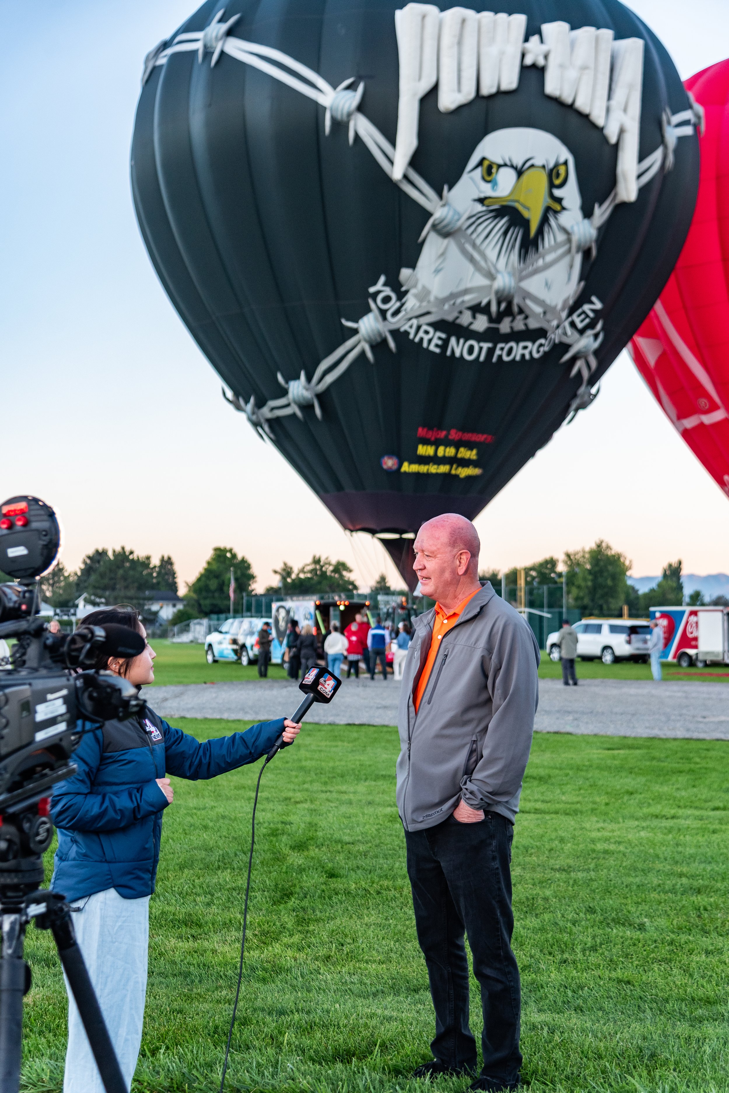 Reporter interviewing a man in front of a large black hot air balloon featuring an eagle and POW/MIA emblem, with "You Are Not Forgotten" text.