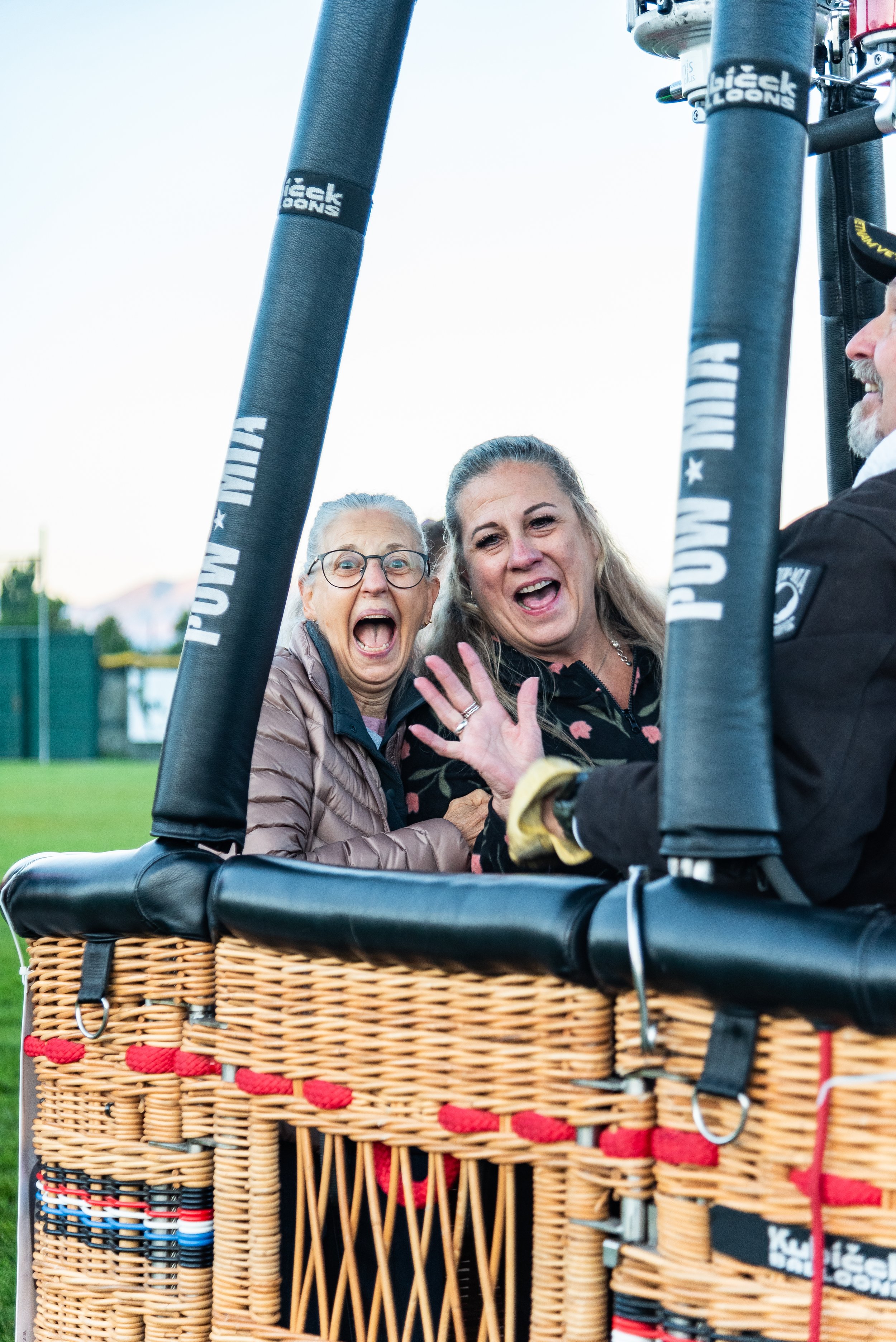 Two women smiling and waving inside a hot air balloon basket on the ground, with a clear sky in the background.