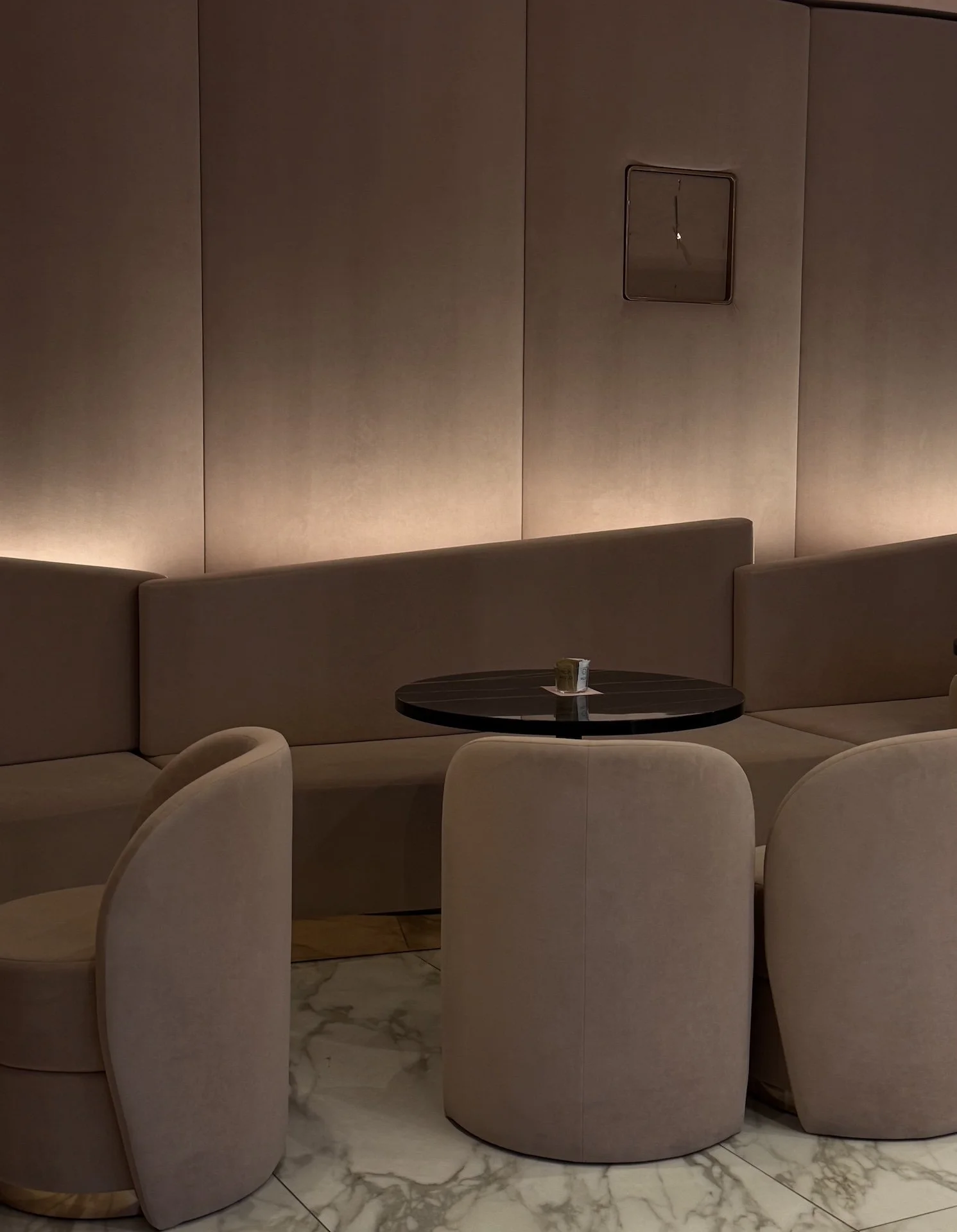 A beige upholstered seating area with a small black round table and three beige chairs, set against a plain beige wall with soft lighting and a small square panel.