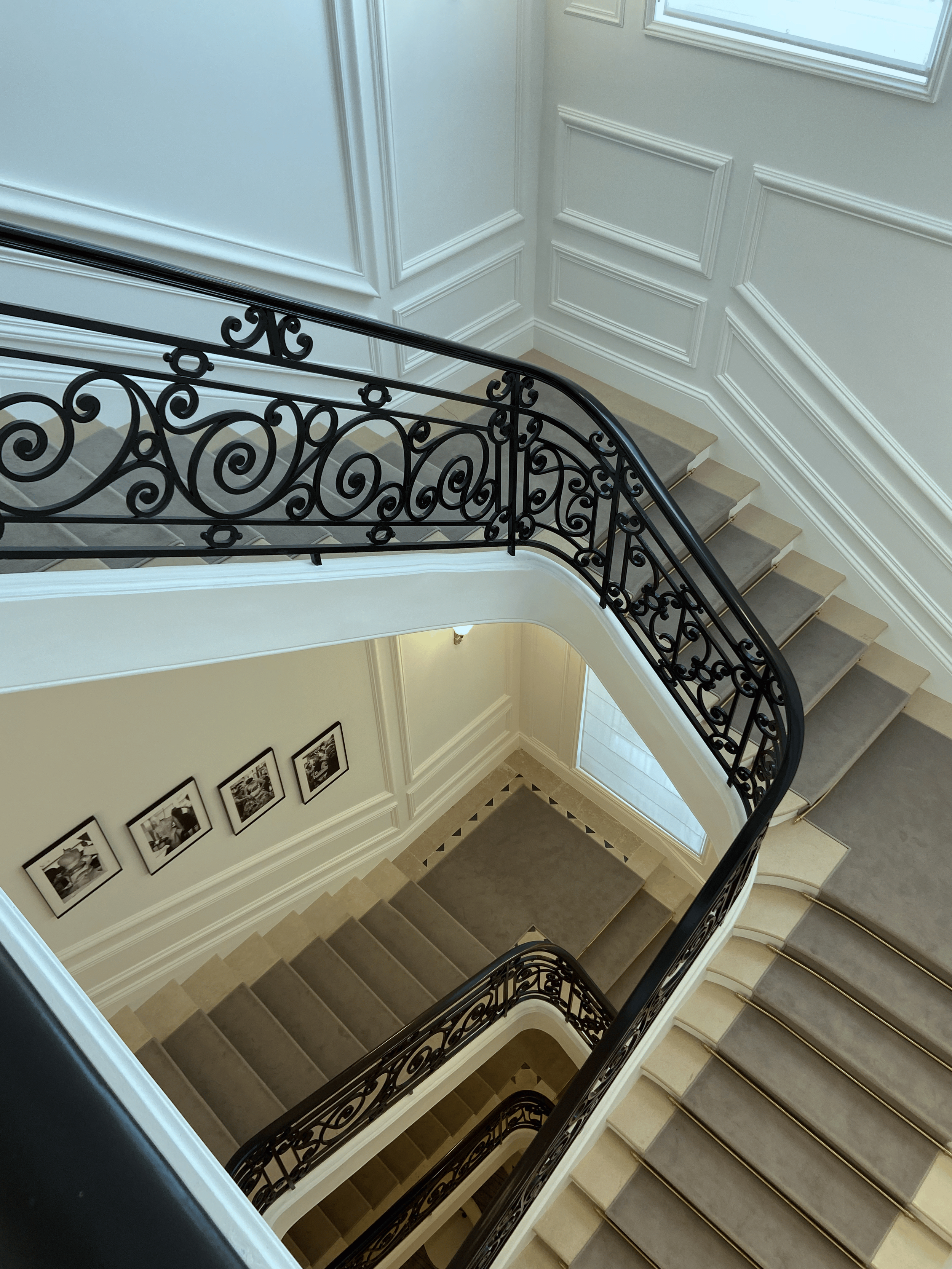 A view looking down a multi-story staircase with ornate black wrought iron railings and beige carpeting on the stairs, surrounded by white wall paneling and framed pictures on the wall.