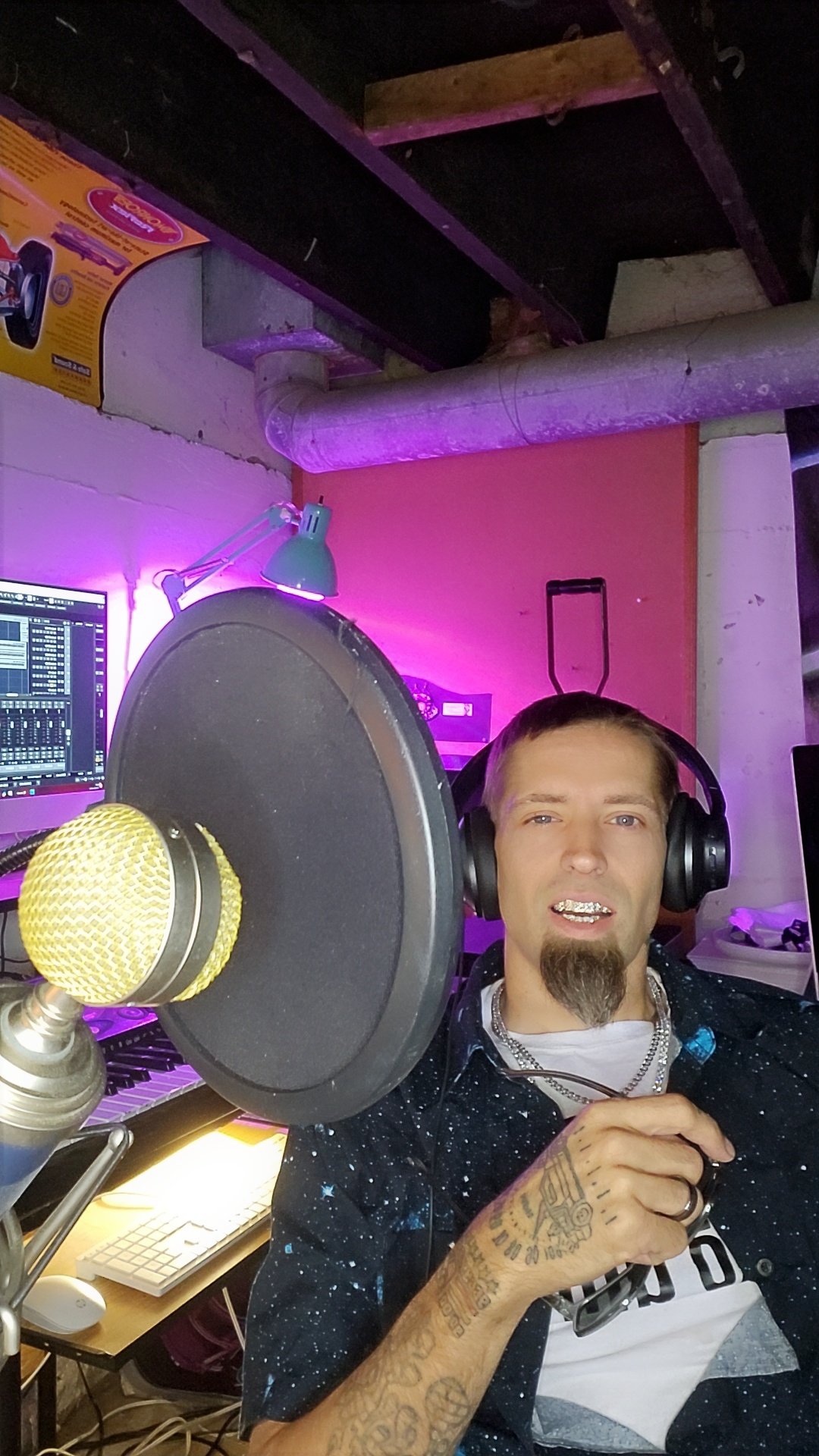 sundawg wearing headphones, sitting in front of a microphone in a music studio with a computer screen and keyboard in the background, illuminated by colorful lighting.