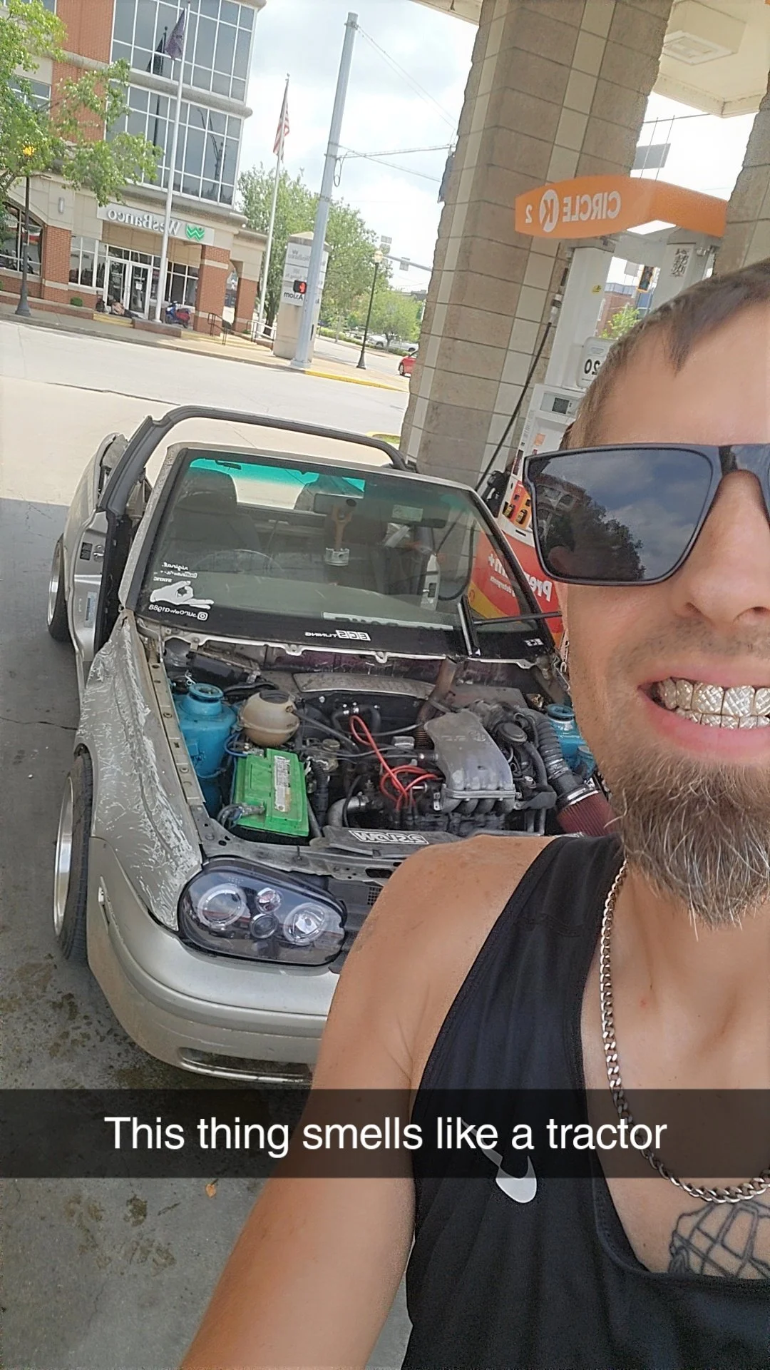 Sundawg w diamond grillz and wearing sunglasses takes a selfie outside a gas station. Behind him, a silver car without a hood is parked at the pump, revealing the engine and various car parts, with the caption 'This thing smells like a tractor'.
