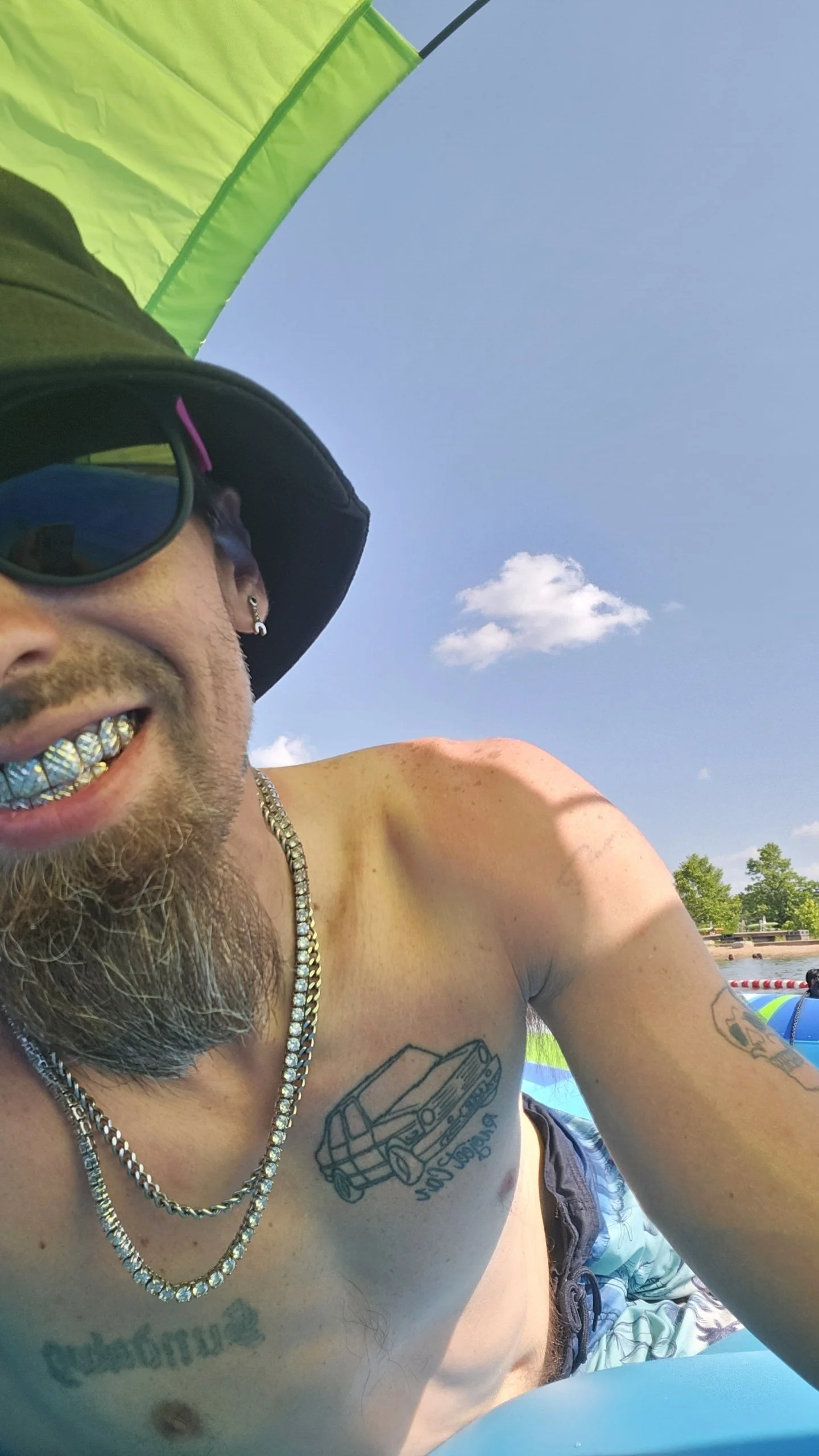 sundawg smiling with gold grillz, wearing sunglasses, a black bucket hat, and jewelry, enjoying a day outdoors with a yellow and green umbrella in the background, and blue sky with a few clouds.