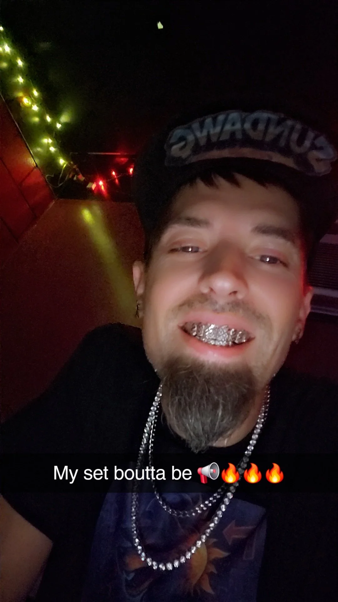 Sundawg smiling with diamond grillz, wearing a black cap with blue letters, and multiple chains around his neck, in a dimly lit room with green and red LED lights.