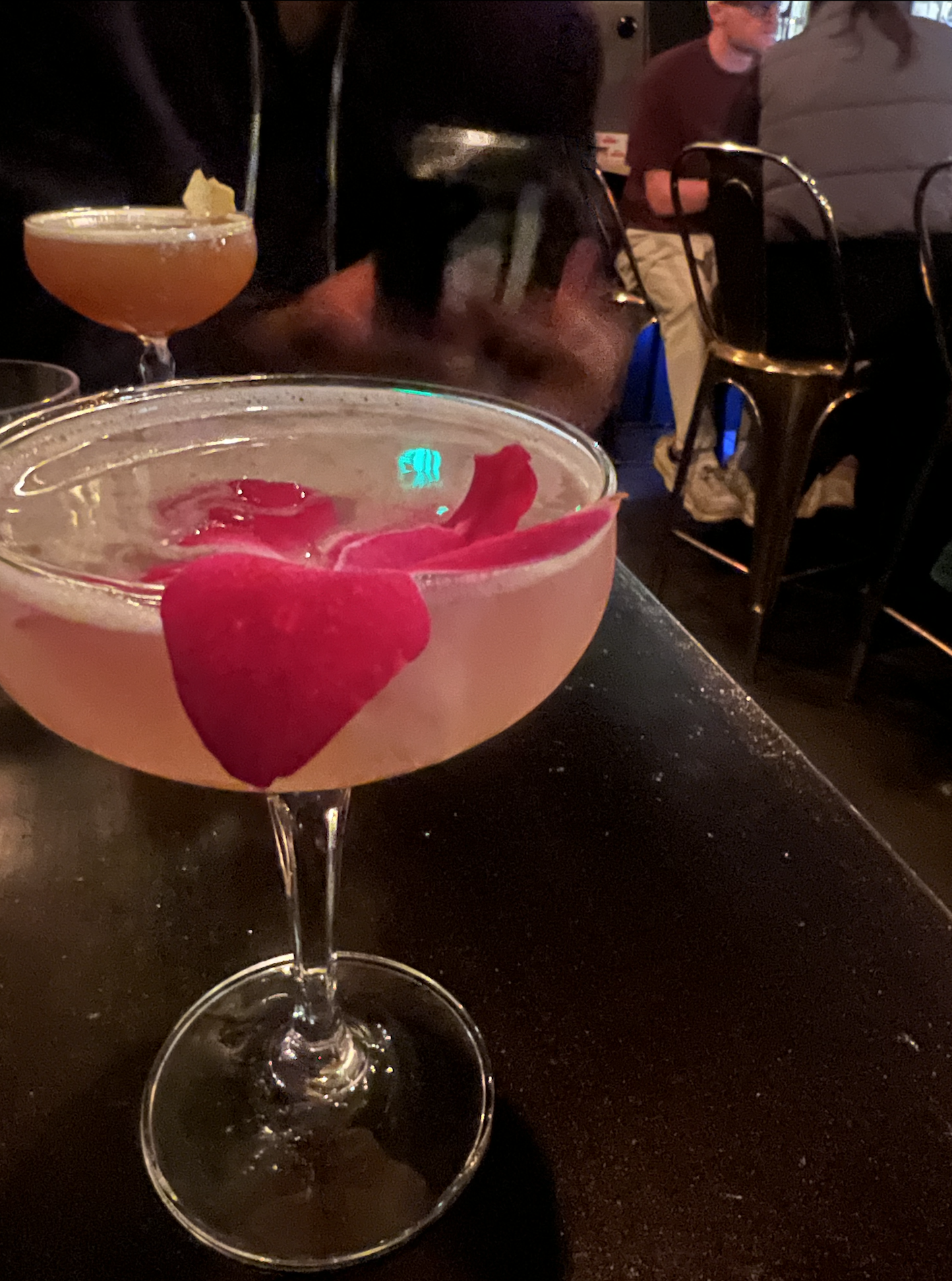 Cocktail in a coupe glass with rose petal garnish on a dark table, person holding a phone in the background.