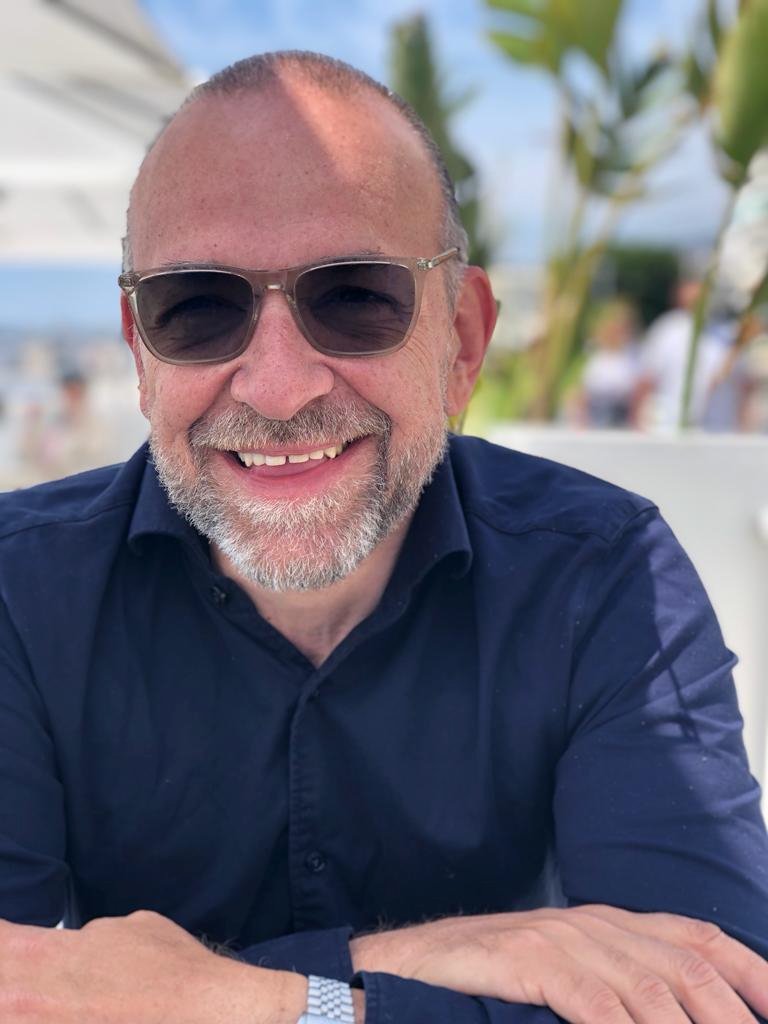 Ein lächelnder Mann mit Bart und Sonnenbrille sitzt an einem Tisch im Freien, im Hintergrund sind Palmen und andere Leute zu sehen.