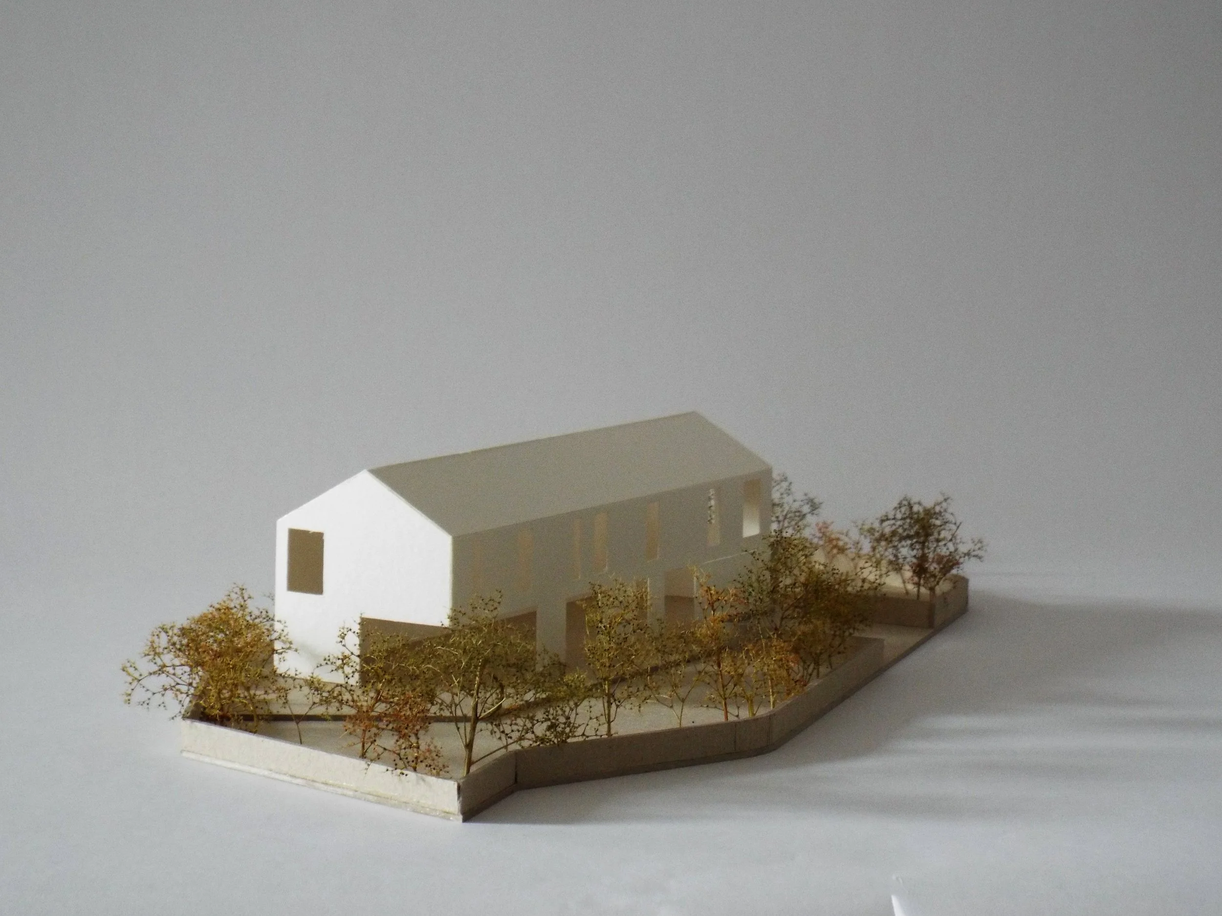 Architectural model of a modern, white building with large windows and a sloped roof, surrounded by trees.