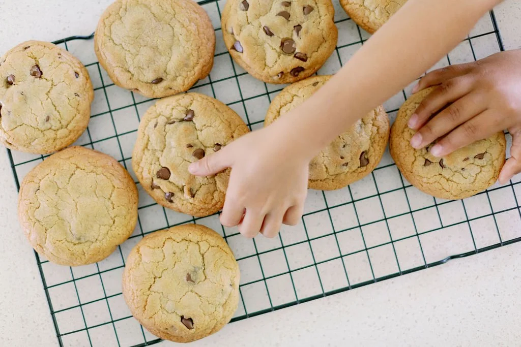 A child’s hands reaching for freshly baked chocolate chip cookies on a wire cooling rack.