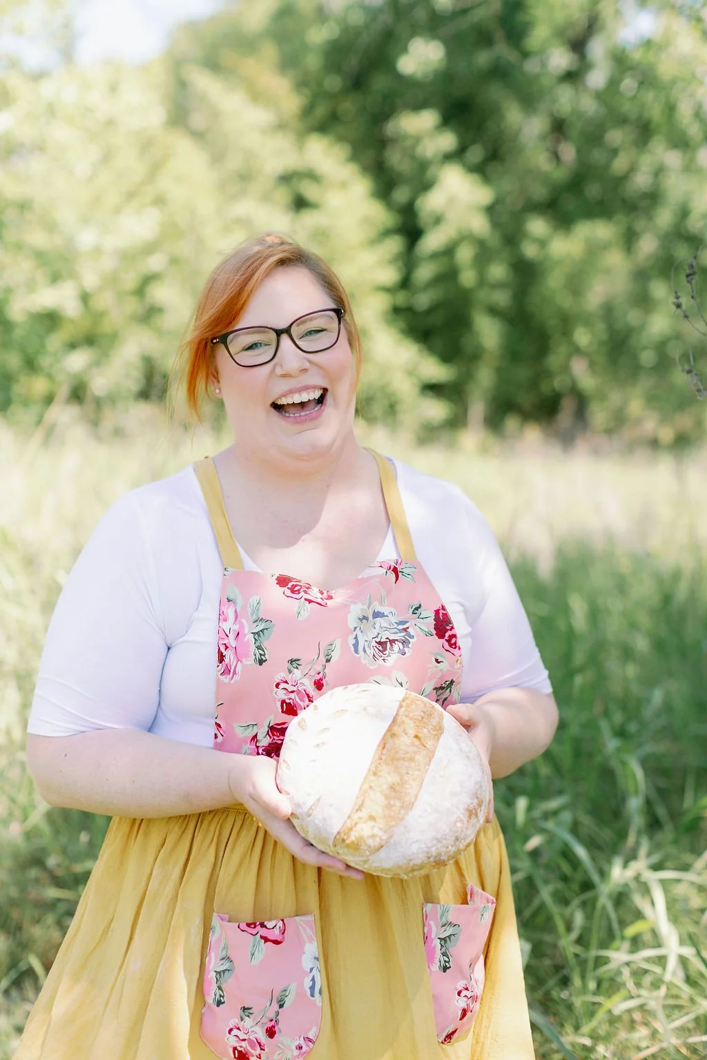 A woman with red hair, glasses, and a big smile is holding a large loaf of bread outdoors in a grassy, wooded area on a sunny day.