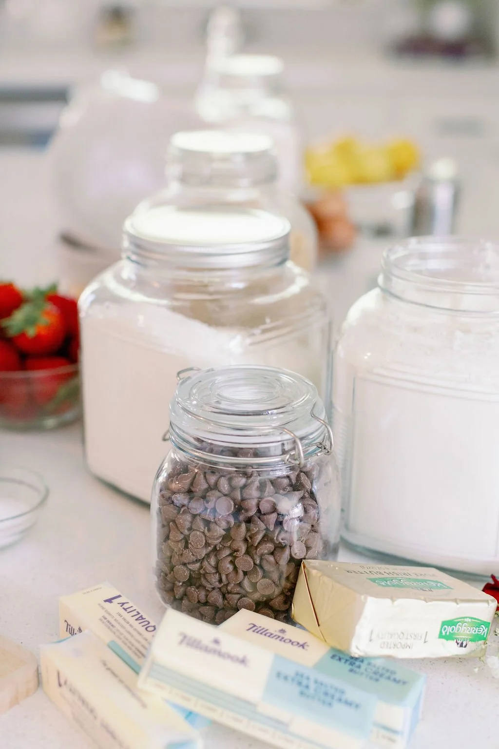 Various kitchen ingredients in glass jars on a countertop, including chocolate chips, flour, sugar, and butter, with strawberries in the background.