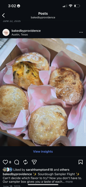 Five large, round sweet rolls or buns in pink polka-dot paper-lined basket, some dusted with powdered sugar, with a golden-brown crust and toppings like corn or fruit pieces.
