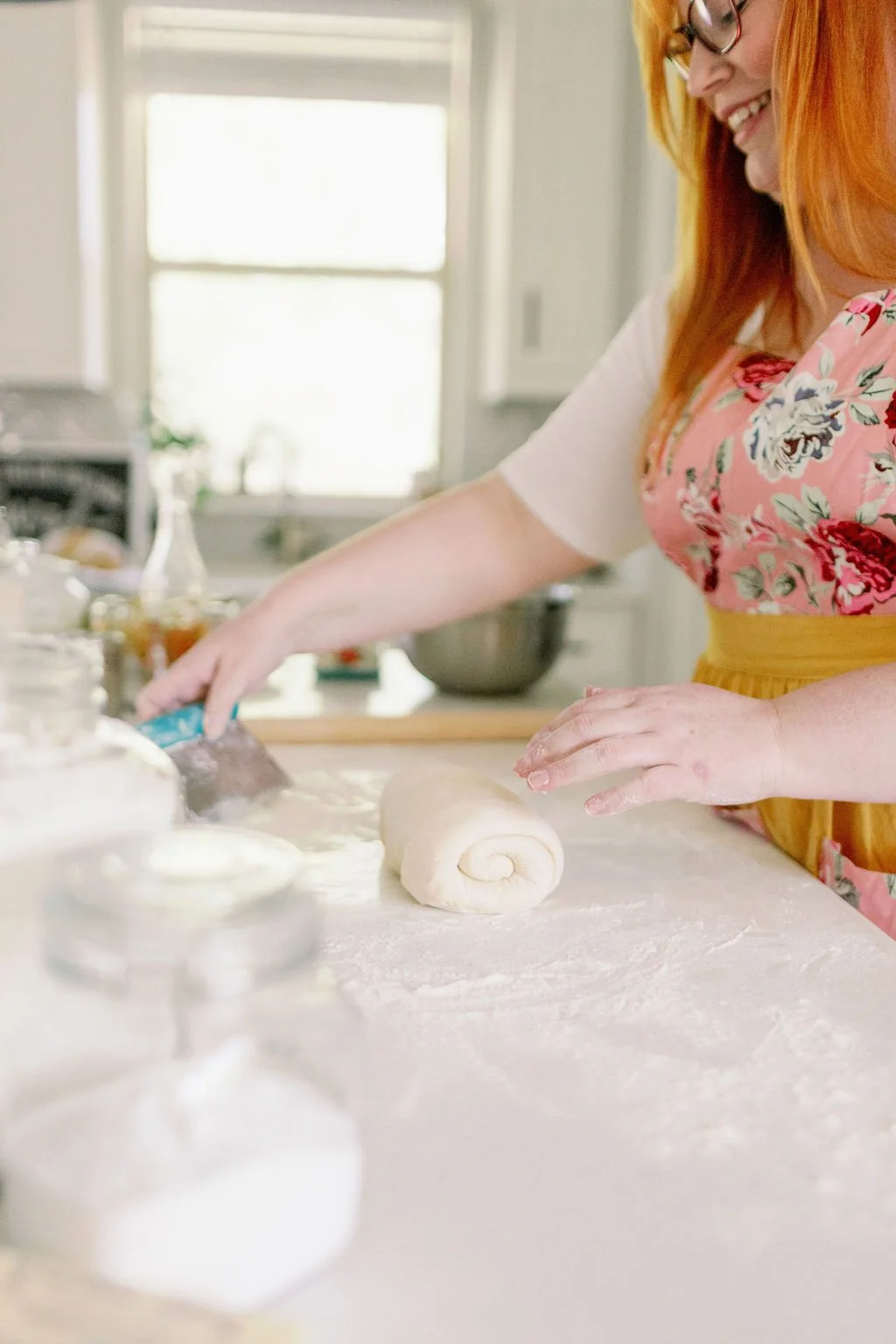 Woman with red hair, glasses, and a floral apron rolling dough on a floured kitchen counter.