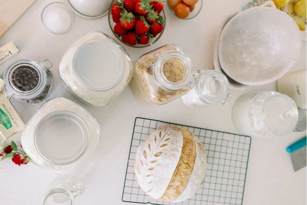 Baking ingredients on a white countertop, including strawberries, eggs, flour, sugar, chocolate chips, milk, and a round loaf of bread on a cooling rack, with a box of butter and a jar of chocolate chips.